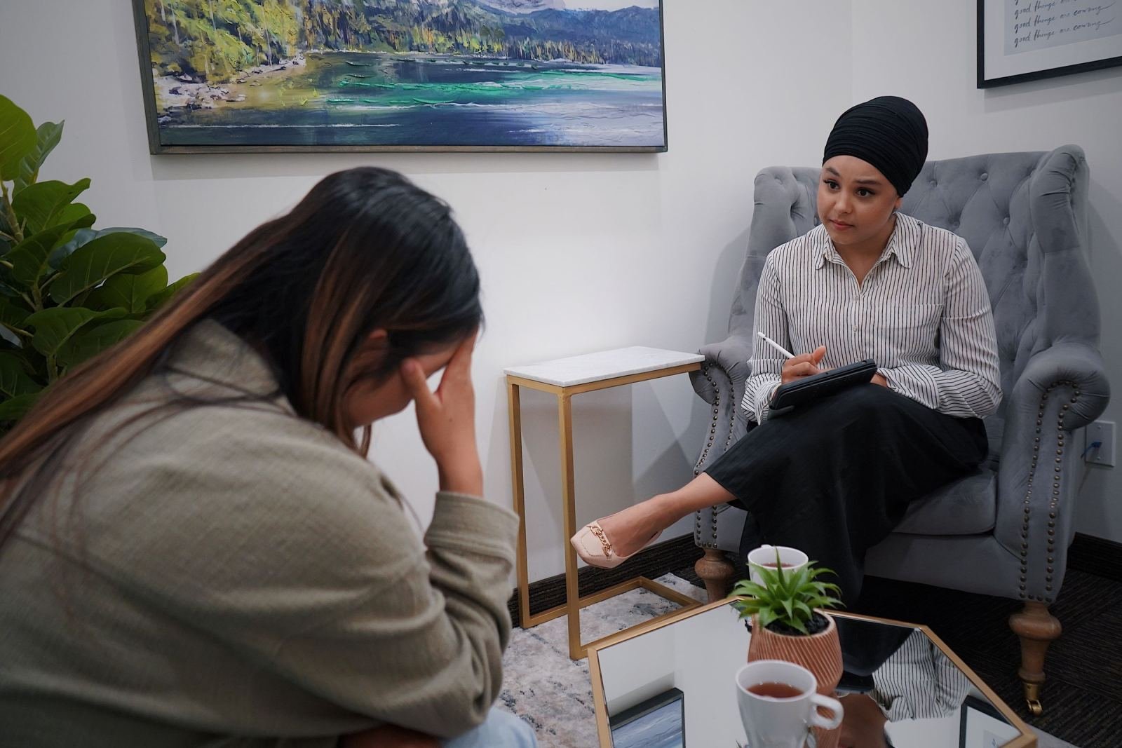 A clinician in a grey armchair speaks with a woman who has her hand to her forehead