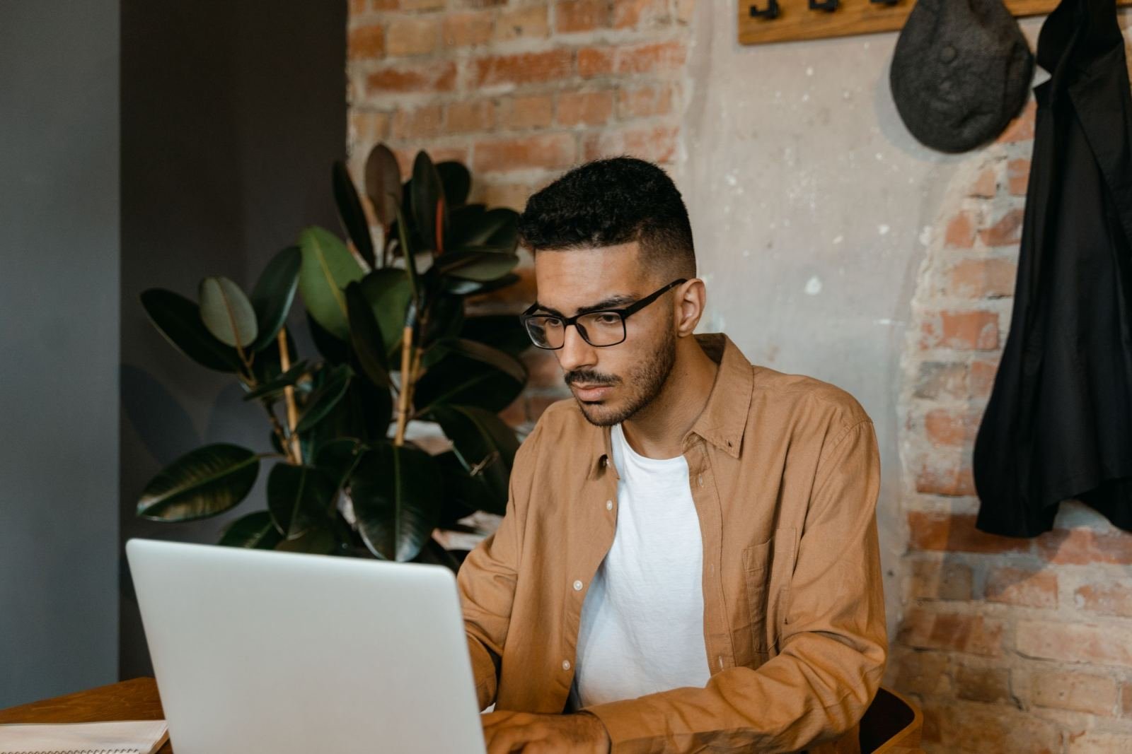 A man wearing glasses views a laptop screen. He sits in a room with a brick wall.
