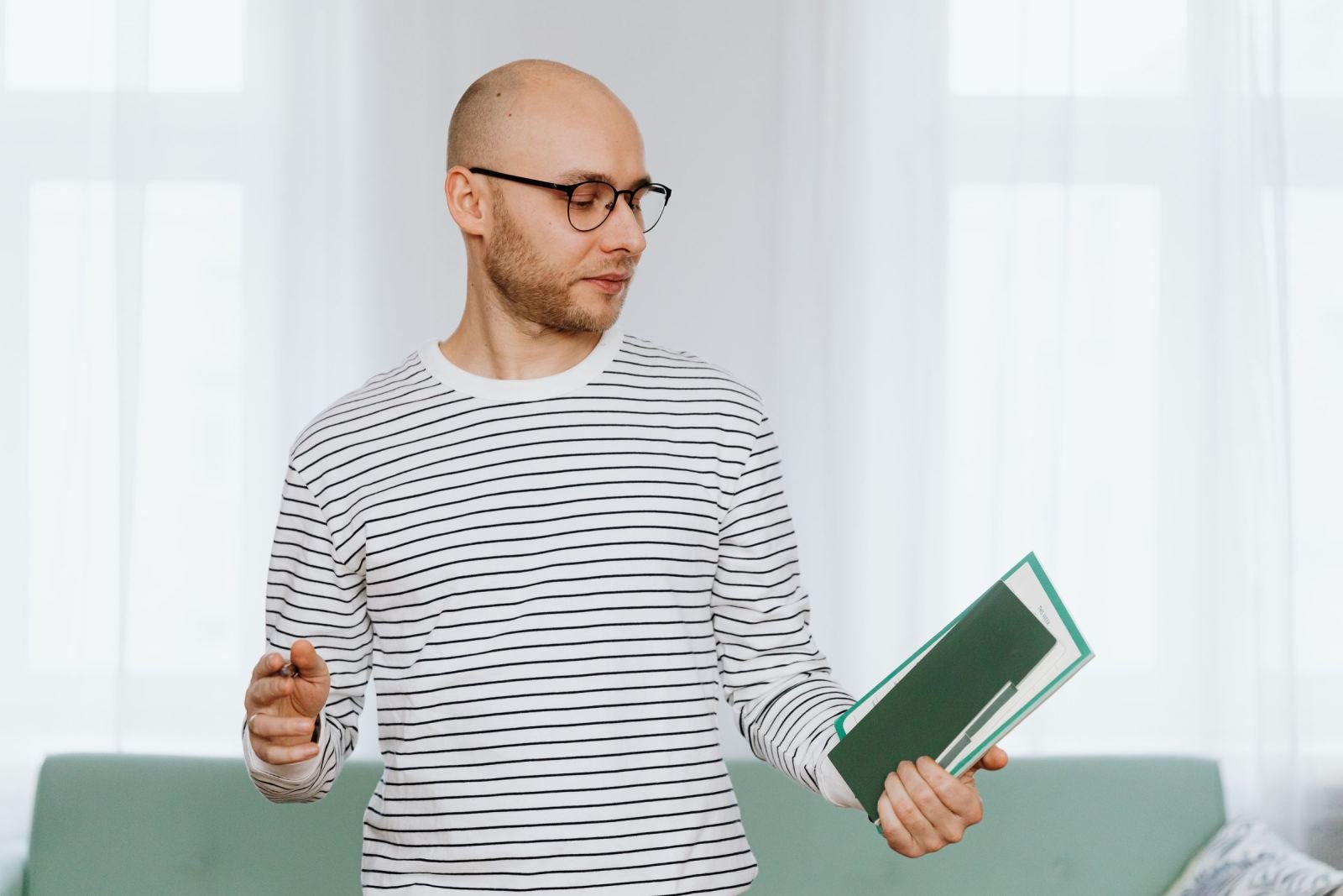 A man with glasses looks at a green notebook in a bright room