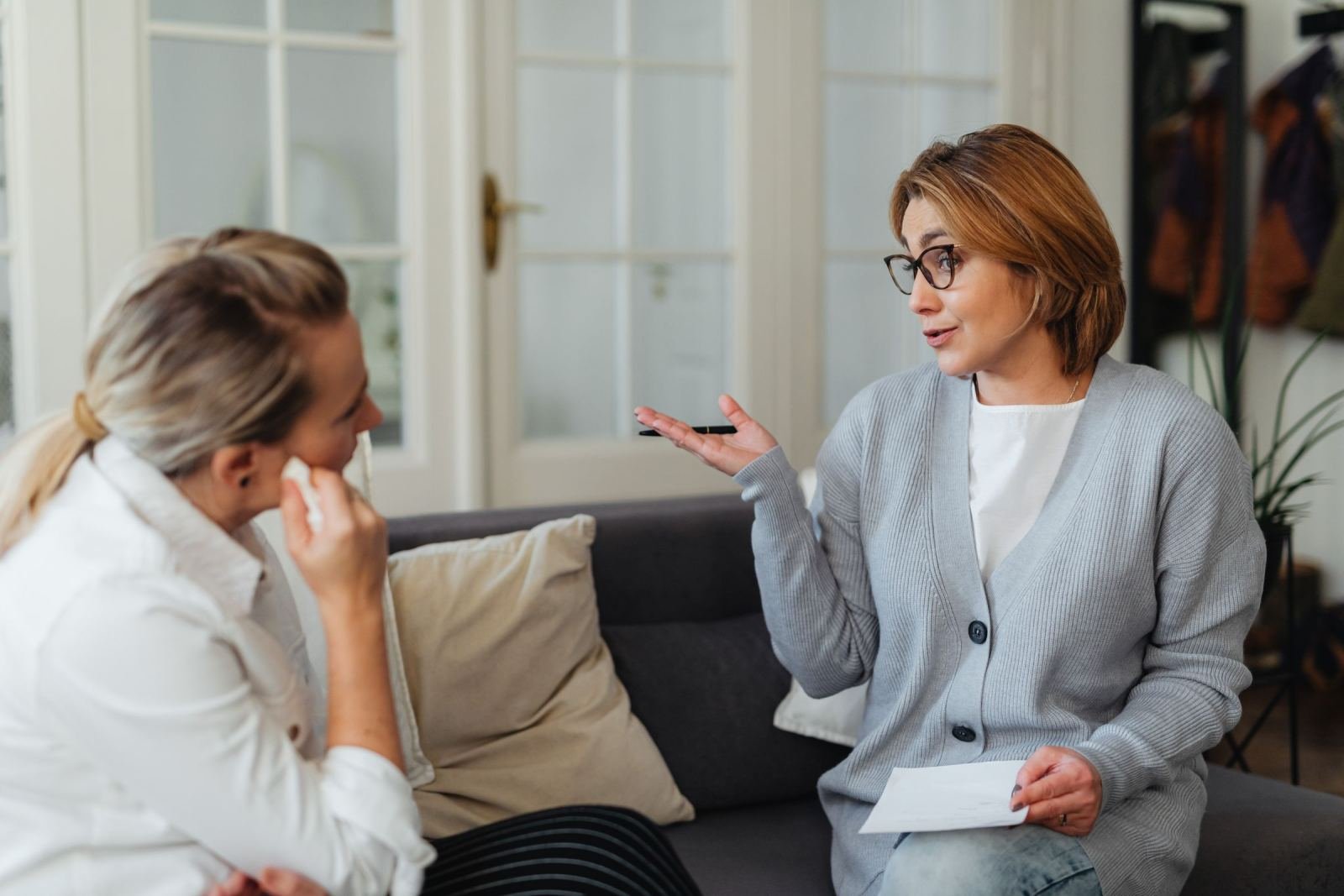 A woman in glasses gestures while speaking to another woman in a bright office