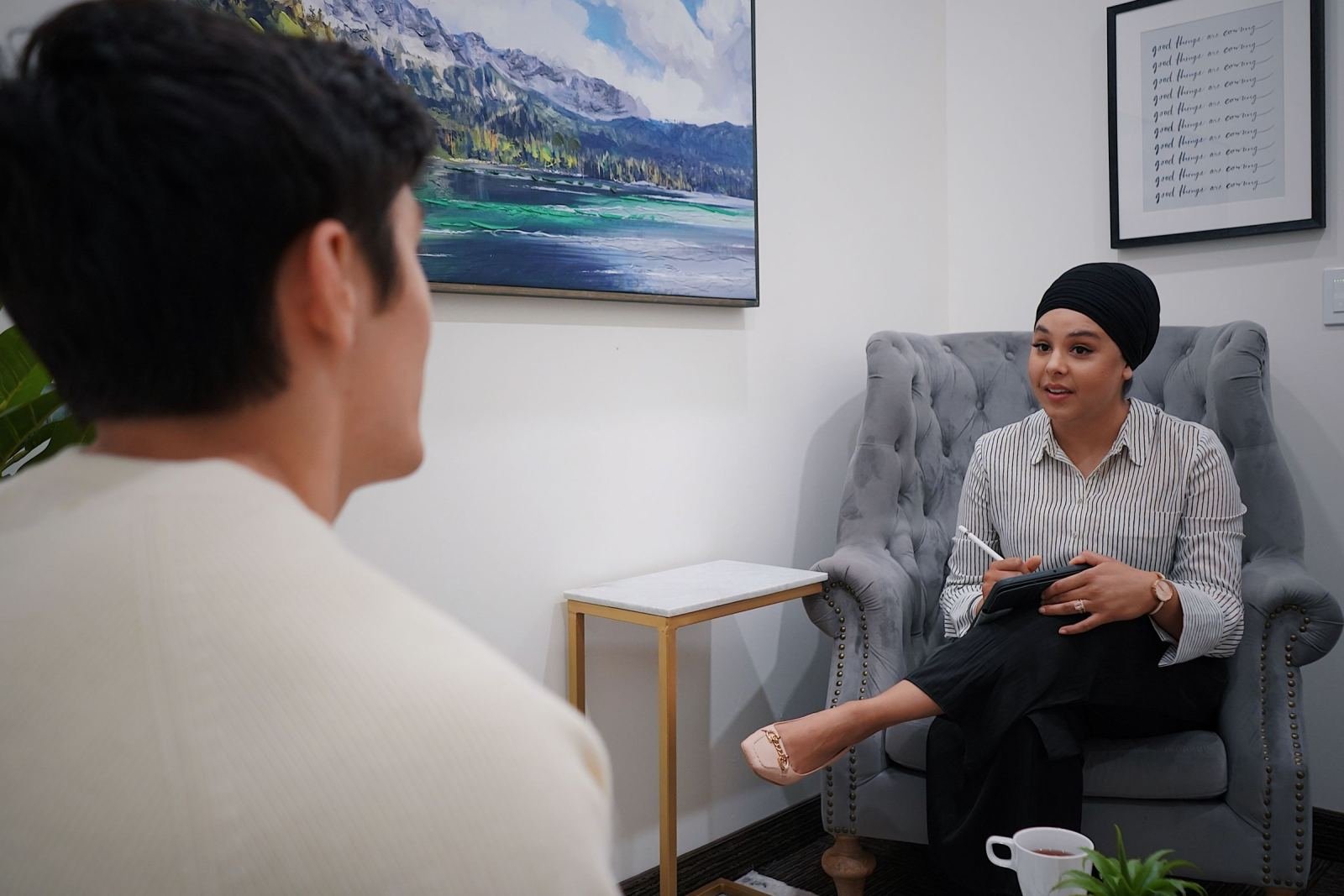 In a therapy room, a woman in a turban speaks with a man while seated in a gray armchair.