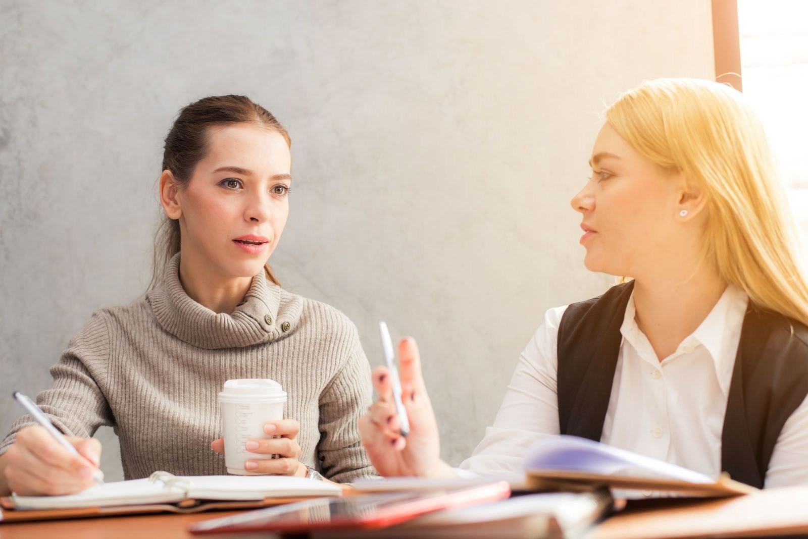 Two women sit at a table. They have notebooks and a coffee cup in front of them.