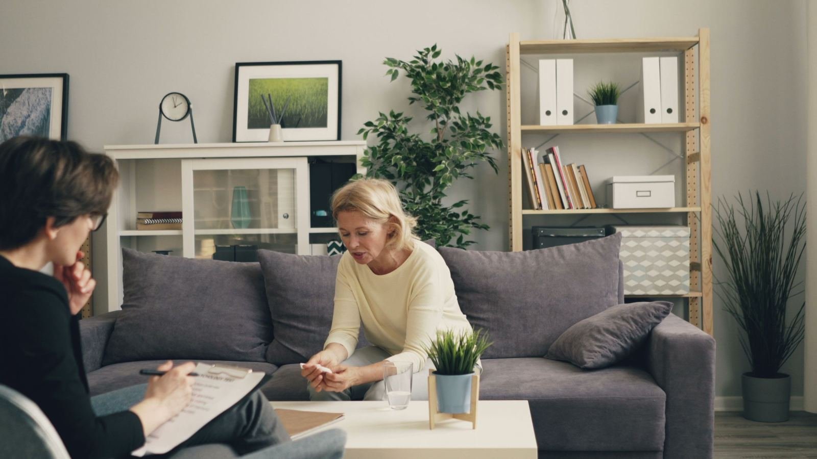 A woman sits on a sofa while a professional with a clipboard listens to her