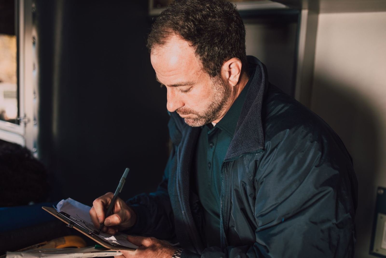 A man in a dark jacket writes on a clipboard at a desk