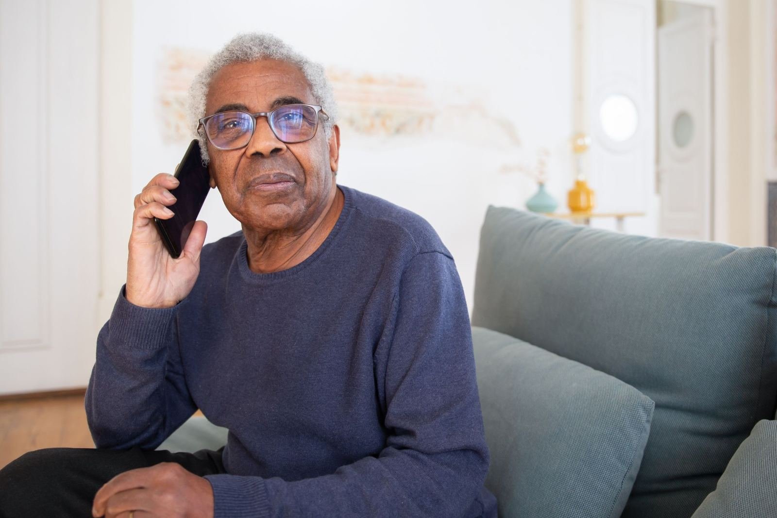 A man with older glasses sits on a couch. He holds a smartphone to his ear.