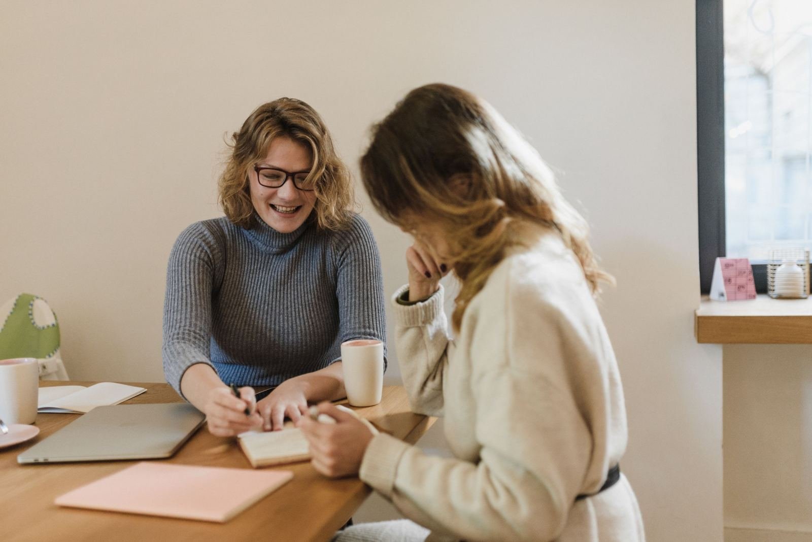 Two women share a table with mugs and notebooks.