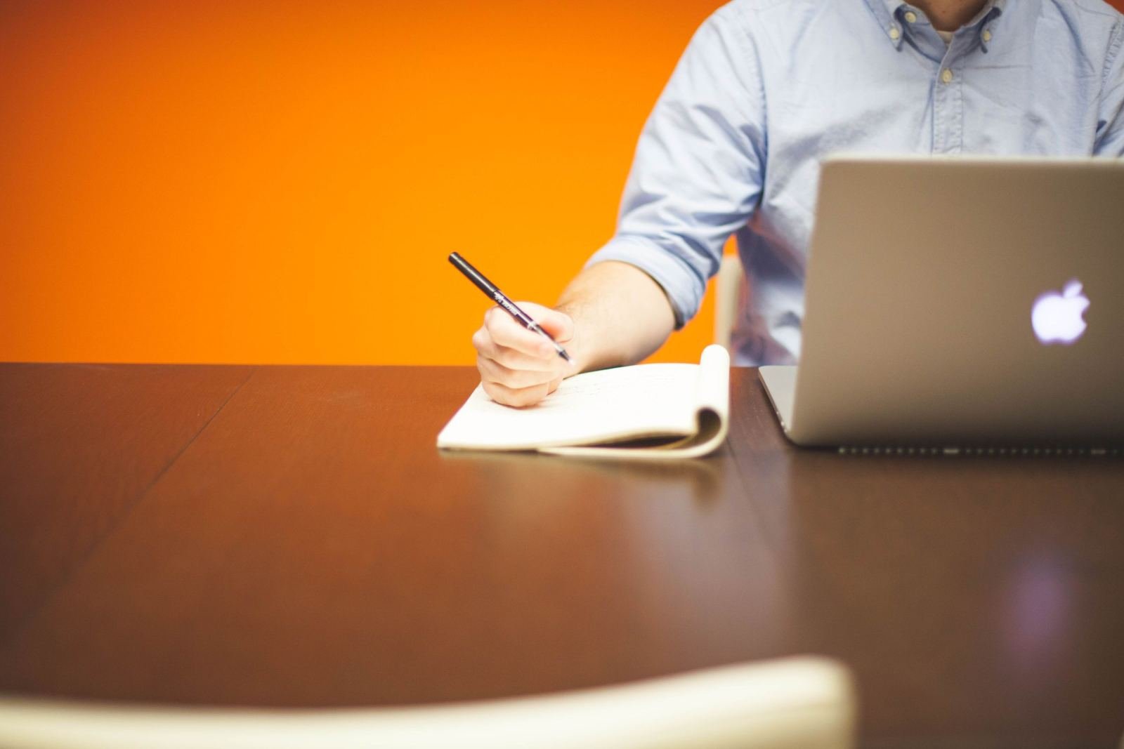 An open laptop sits on a wood table. Beside it, someone writes in a notebook.