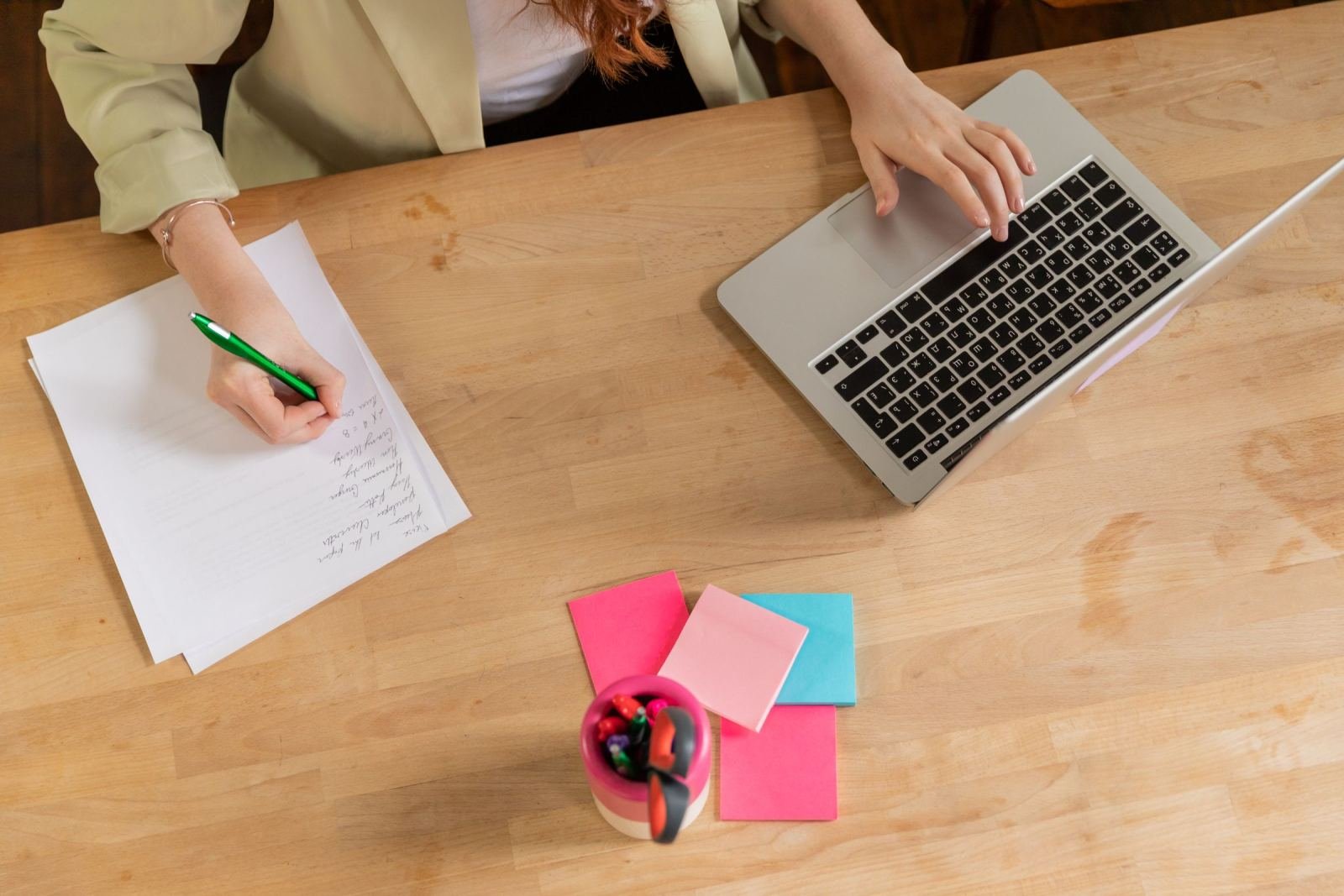 Someone in a blazer makes a list on paper beside a laptop.