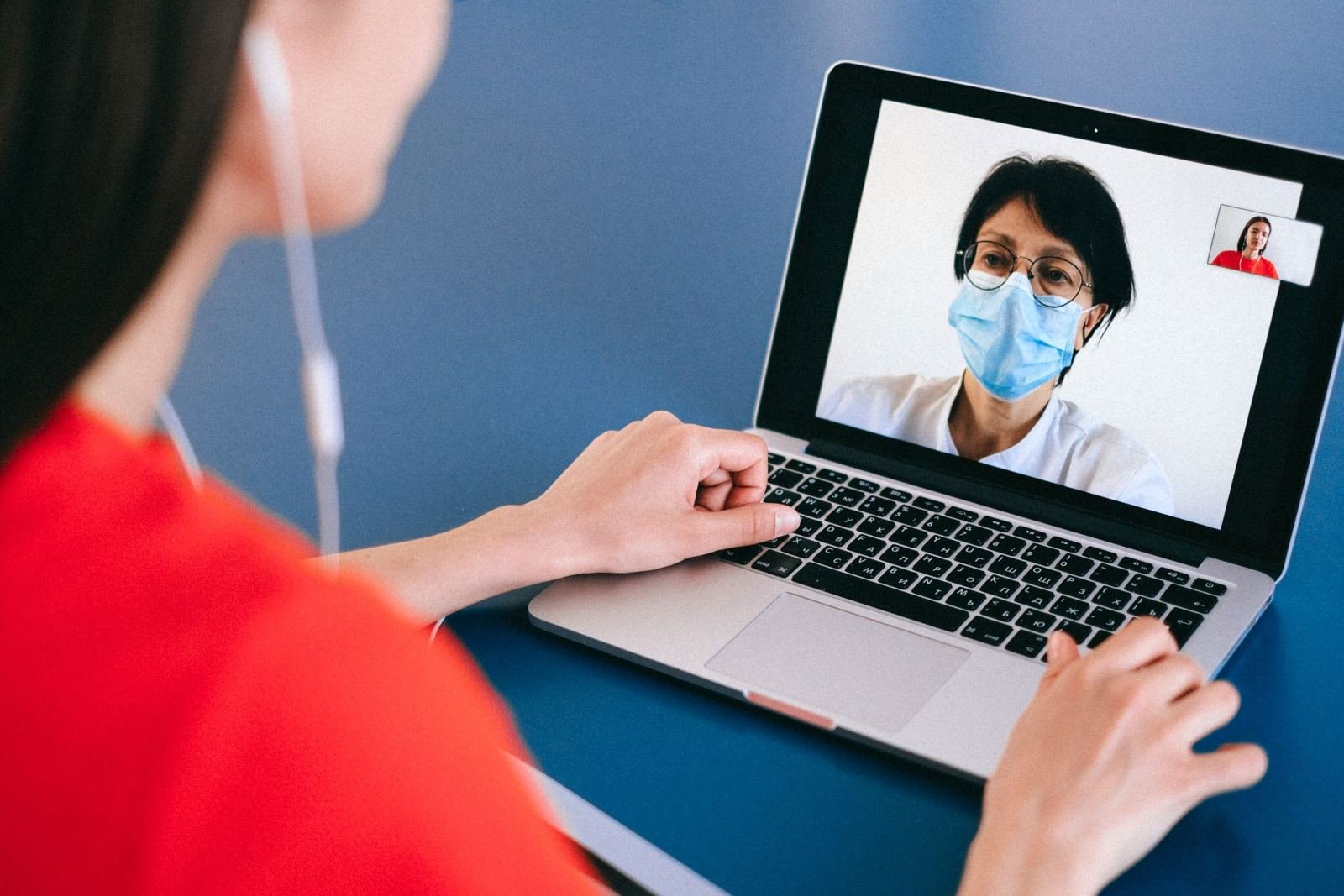 A person in a red shirt talks to a masked healthcare provider on a laptop via video call.
