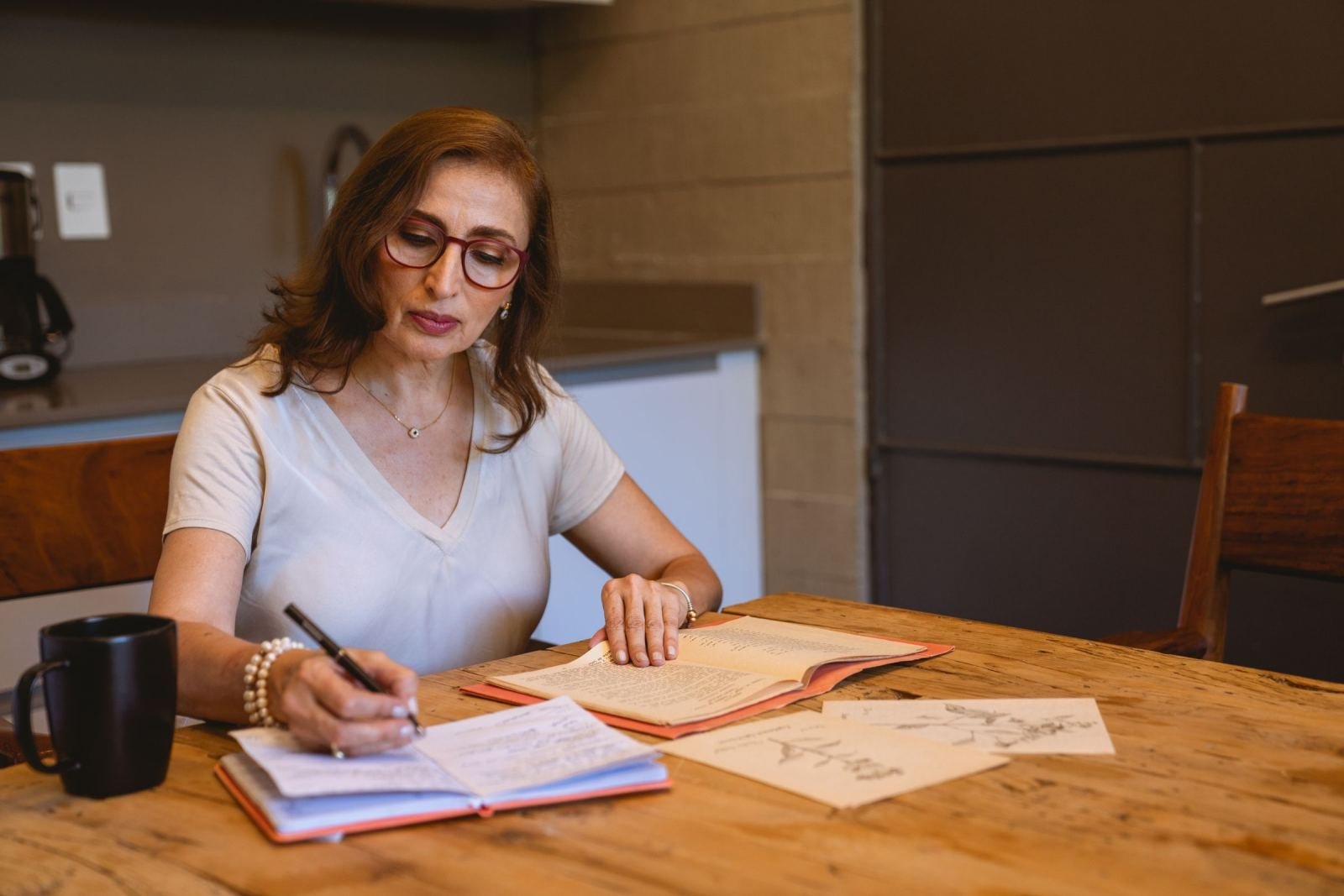 A woman wearing glasses writes notes in a book on a wood desk.