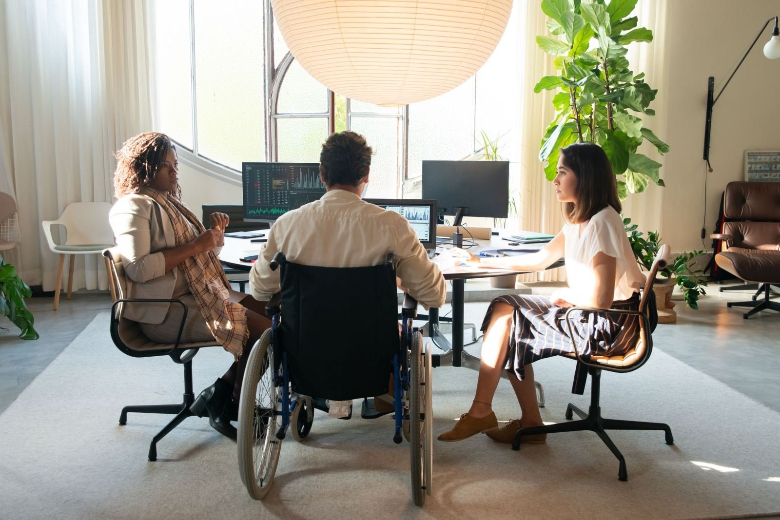 A person in a wheelchair and two women talk around a desk with computer monitors