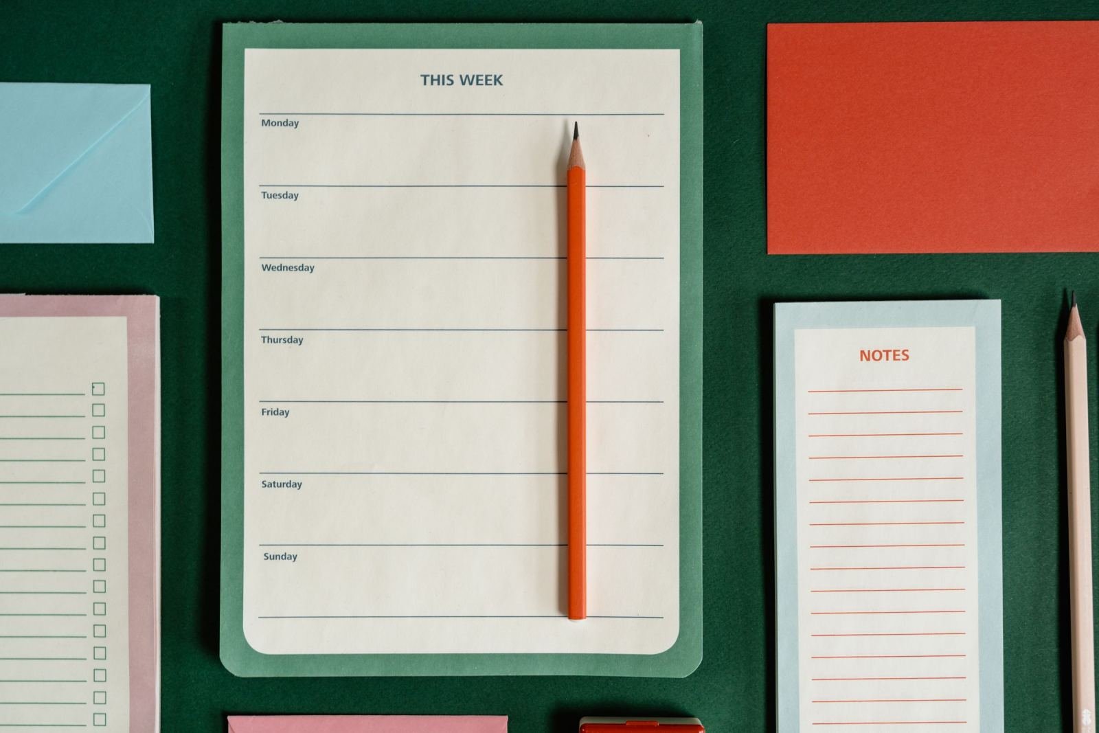 A weekly planner and orange pencil on a green surface with colored notes