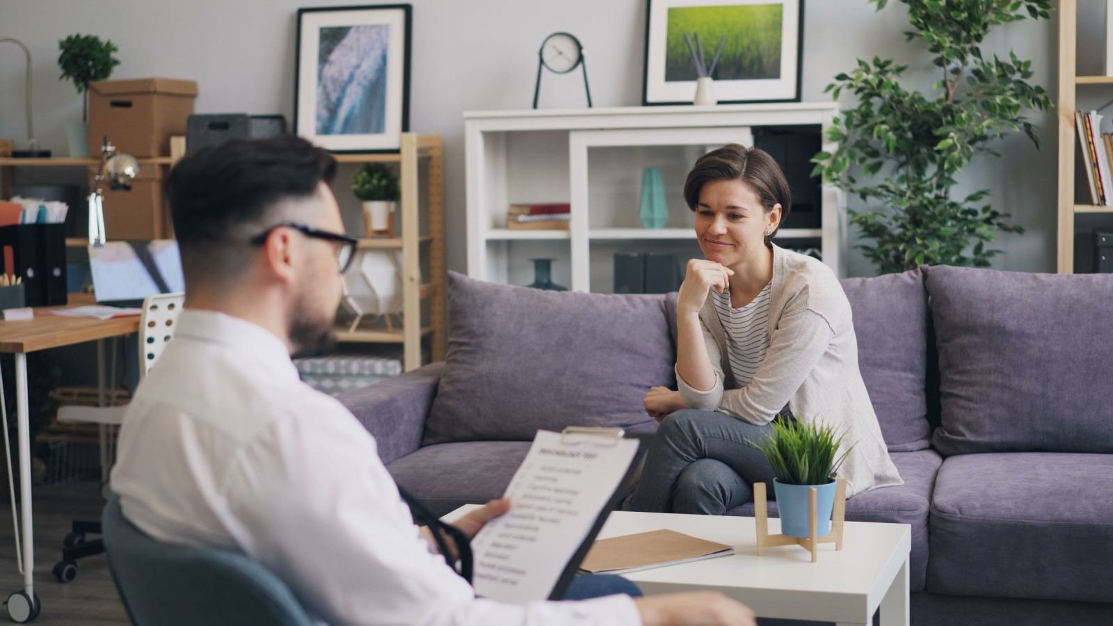 A man with a clipboard speaks with a woman sitting on a purple sofa in a bright office