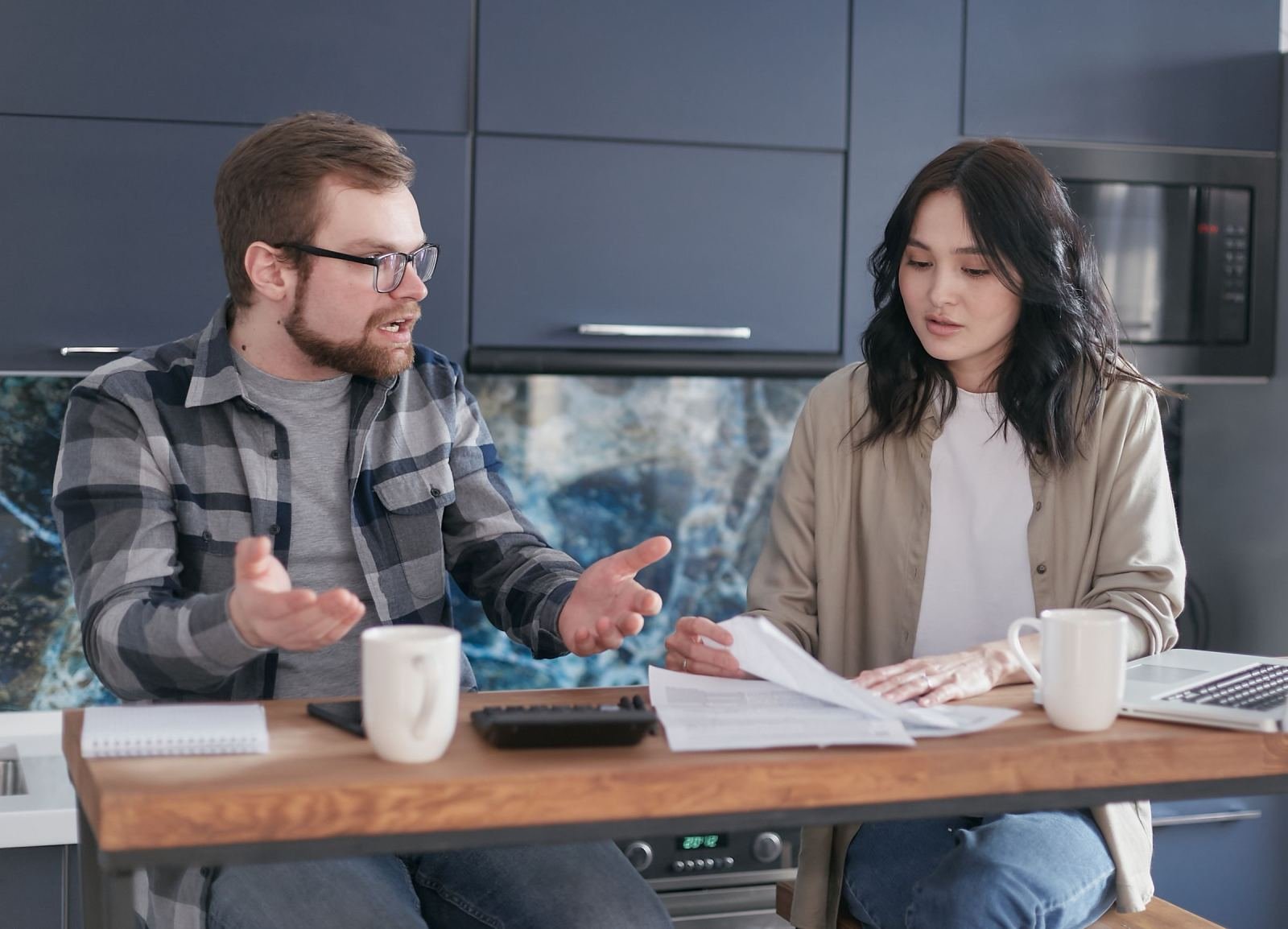 A man and woman sit at a wooden table, looking over papers and using a calculator.