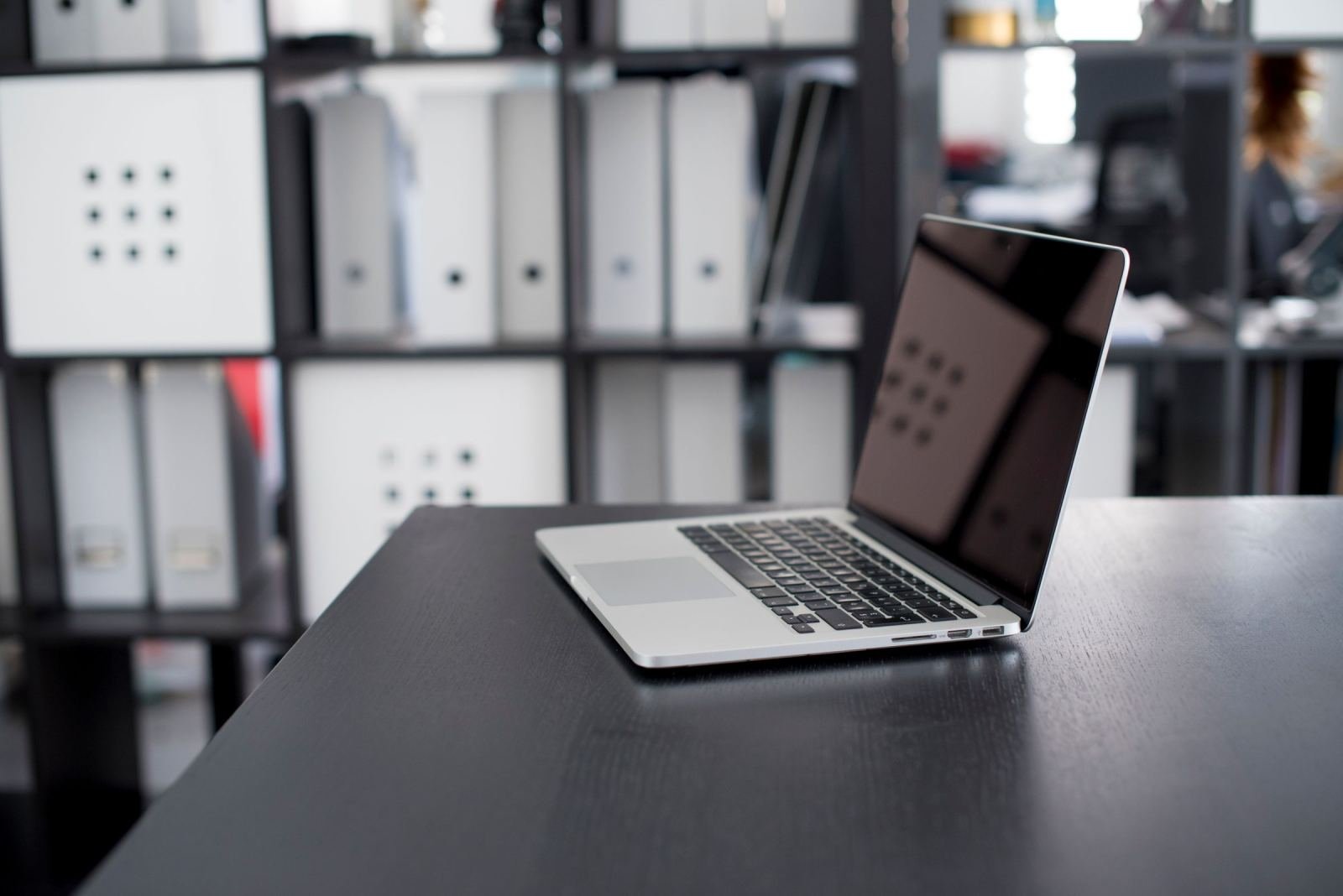 A dark desk holds an open laptop. Blurred office shelves sit in the back.