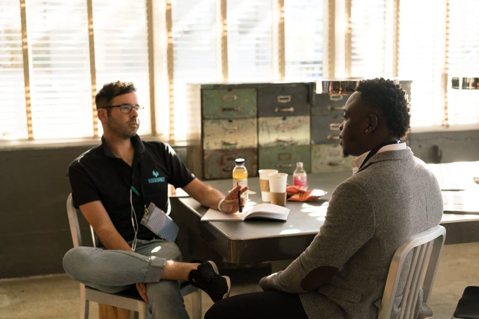 In a bright office, two men talk at a table while going over a notebook.
