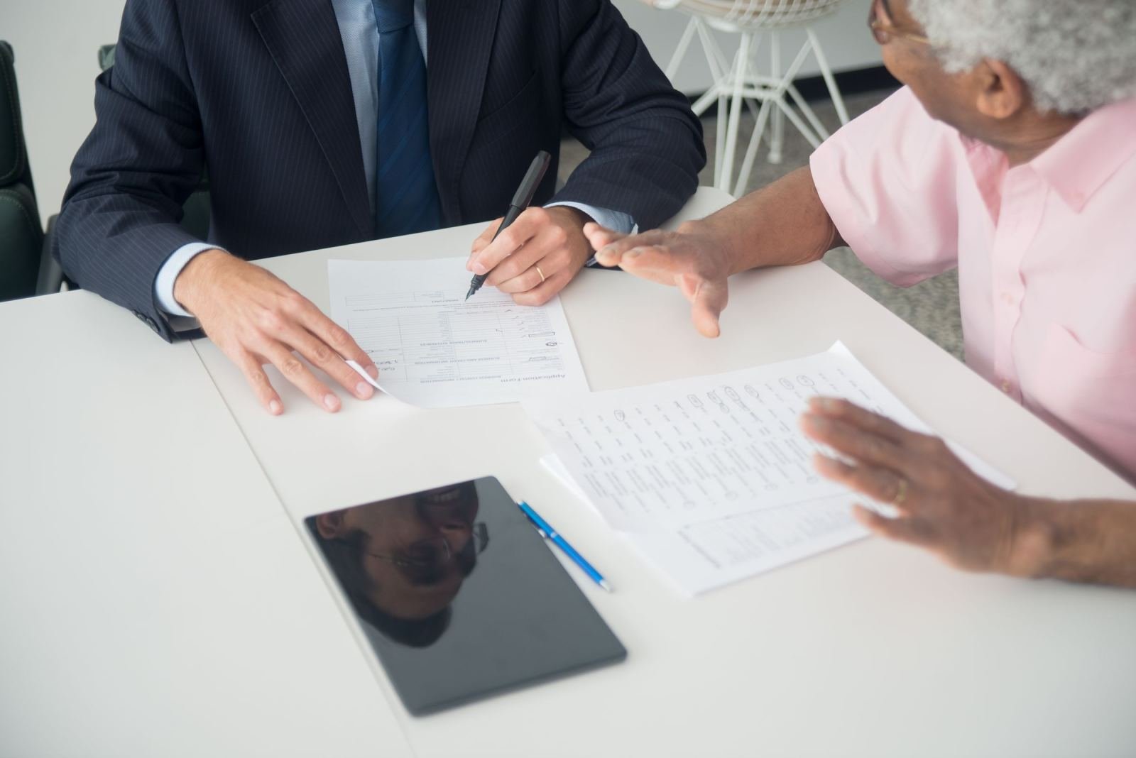 A man in a pink shirt sits across from a professional in a suit. They talk at a desk covered in papers.