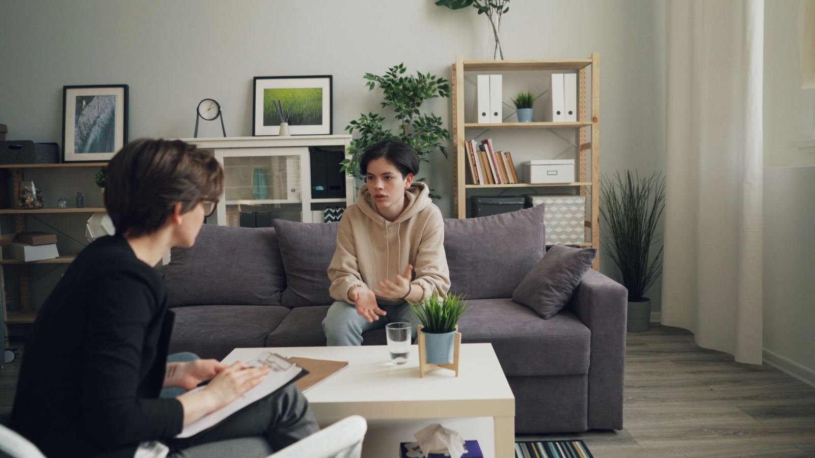 A clinician in a black blazer sits on a grey sofa and talks with a teenager.