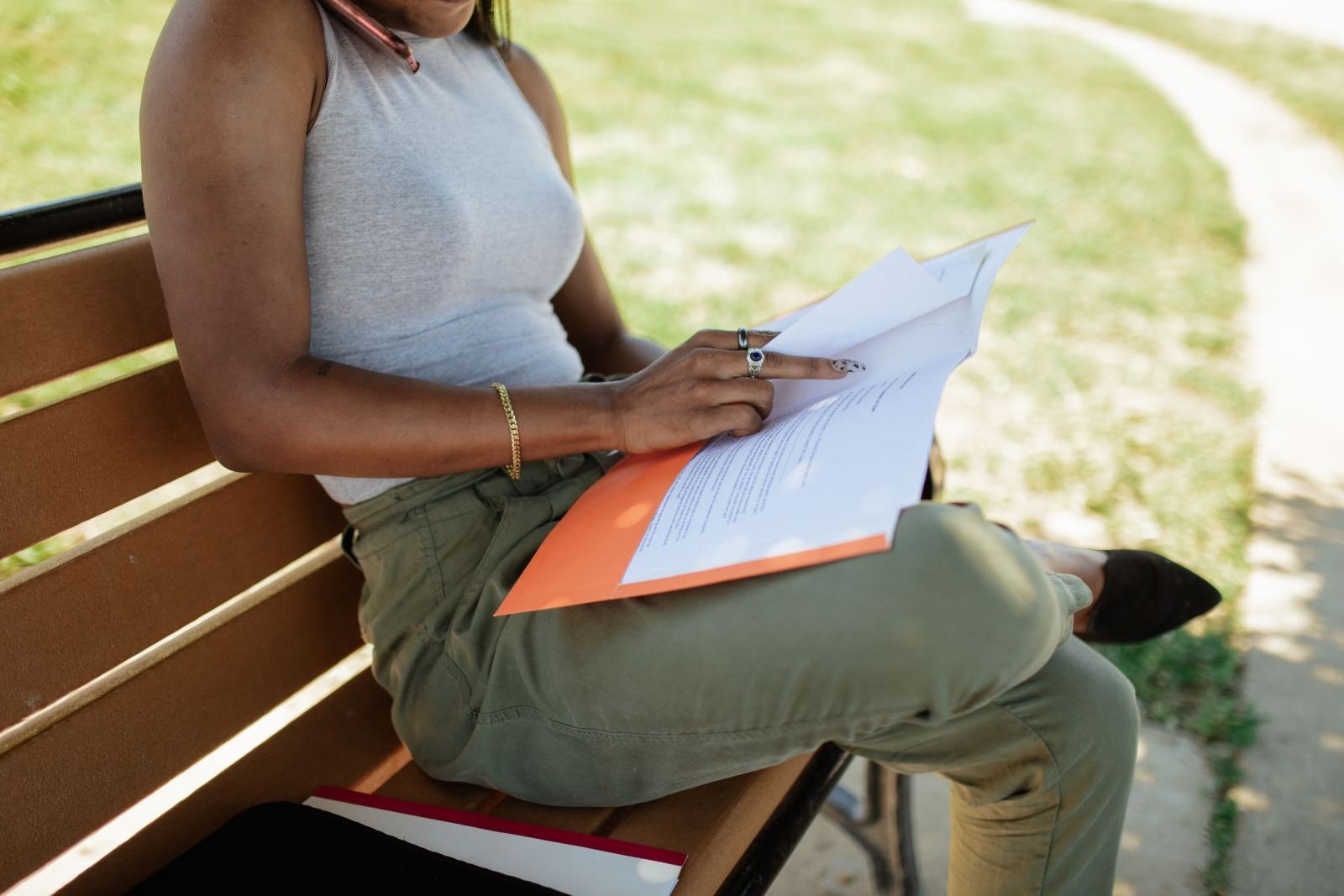 A person sits on a park bench holding a paper document that has an orange cover.