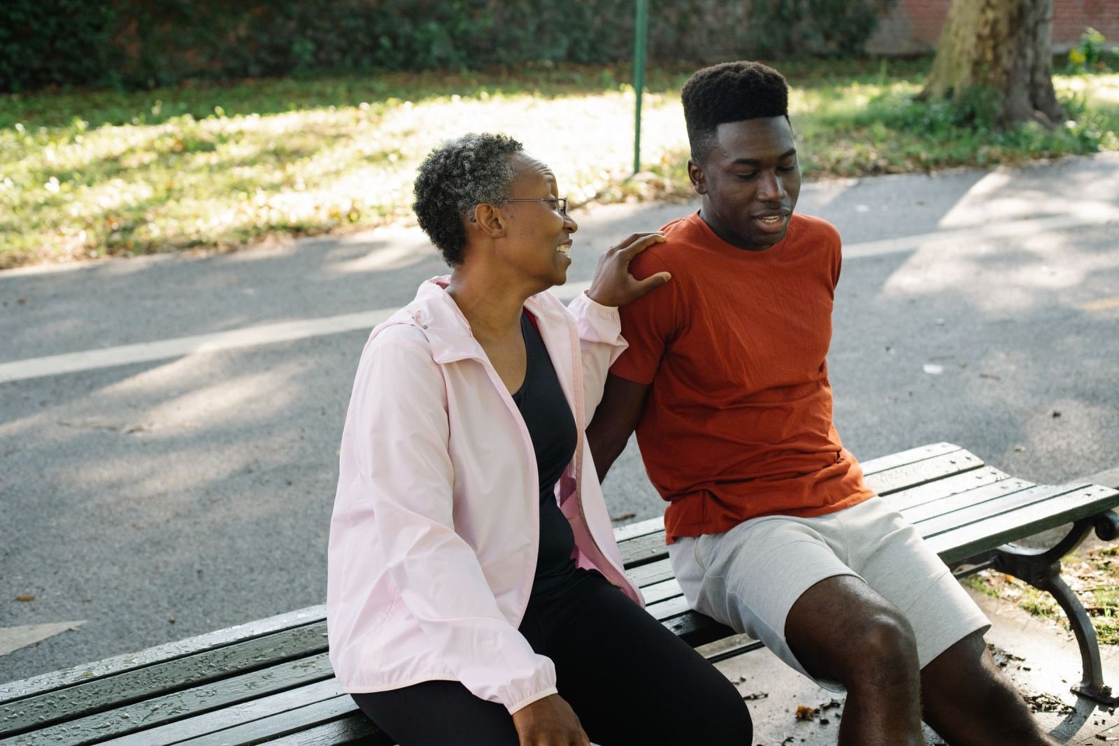 A park bench holds a young man and an older woman, who rests her hand on his shoulder.