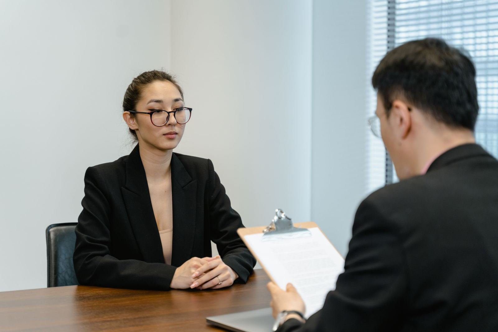 A man with a clipboard talks to a woman in a black blazer at a desk