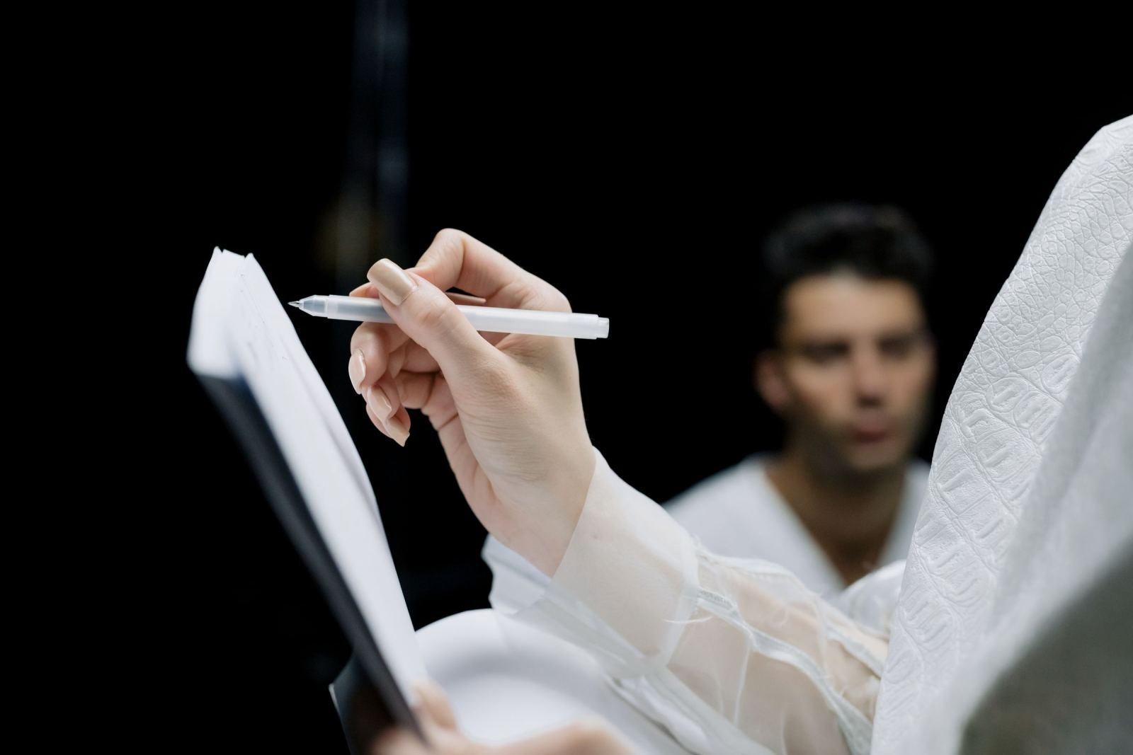 A person writes in a notebook while a man stands out of focus behind them.