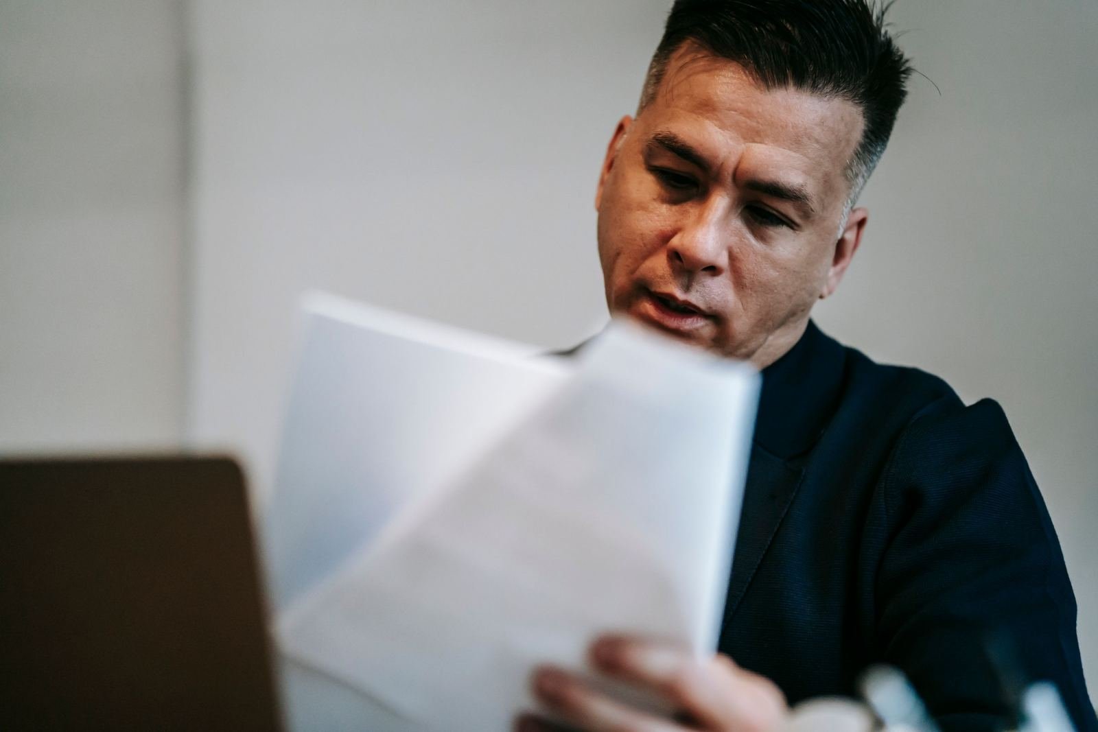 A man reads a white document while sitting at a desk with a laptop
