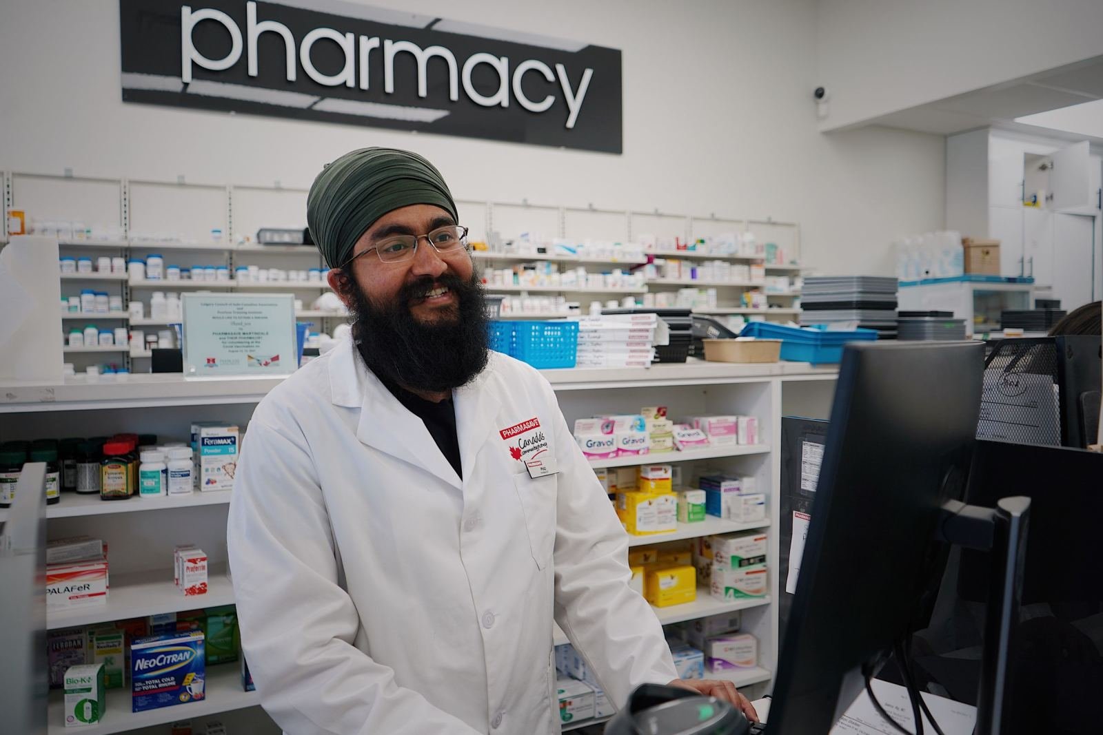 A happy pharmacist wearing a white coat and a green turban sits behind a counter.