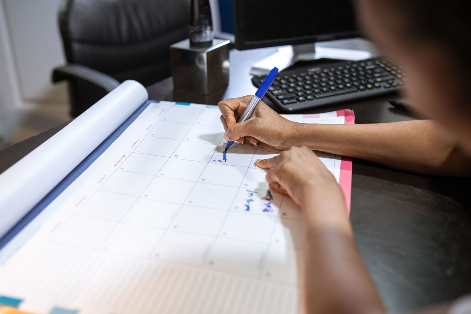 A person writes on a large paper calendar at a desk with a computer