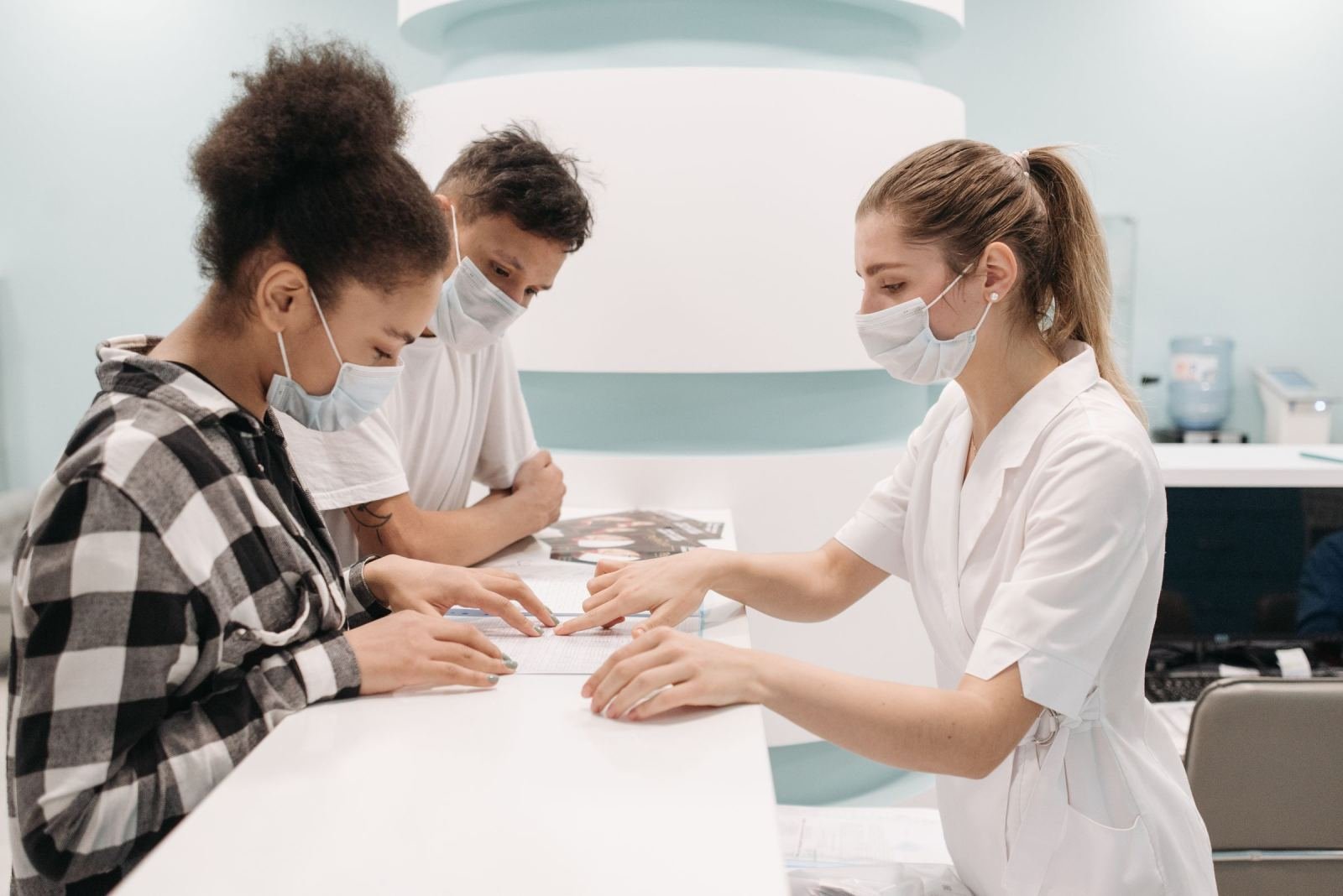 A masked clinician in a white coat looks over papers with two people at a counter