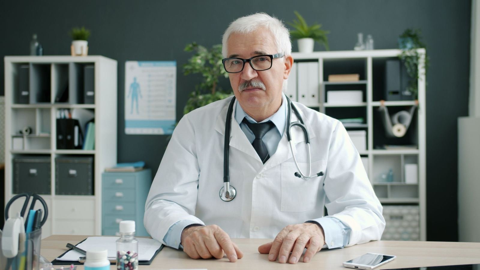 A doctor wearing a white coat and stethoscope sits at his desk.