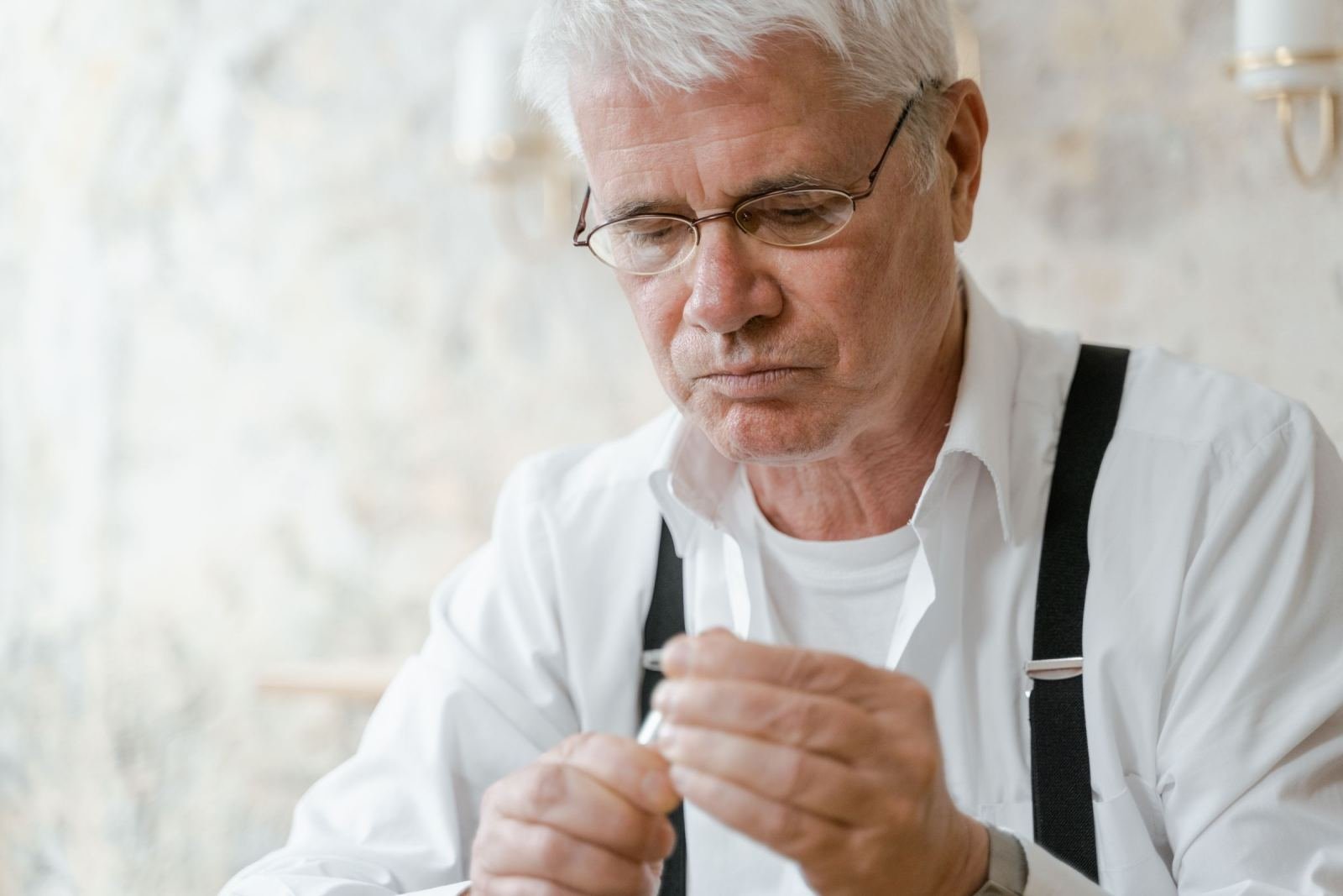 A senior man wearing glasses and suspenders stares at a thing he holds.
