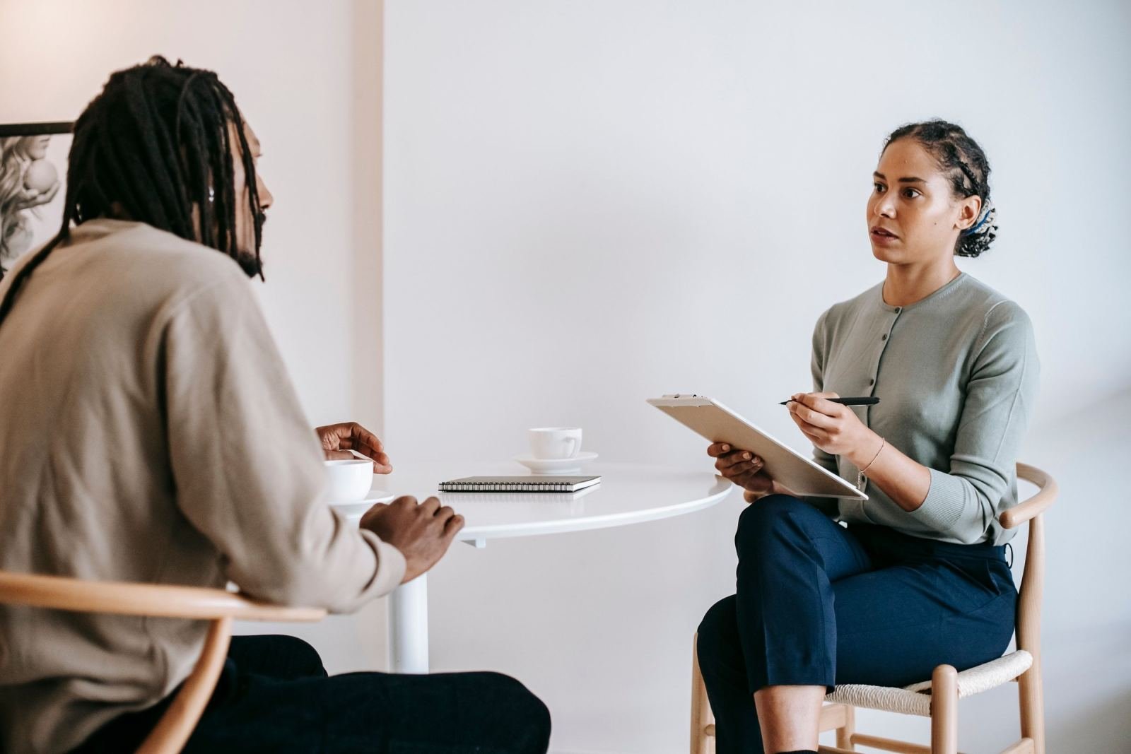 A man sits at a small white table. He talks with a counselor who holds a clipboard.