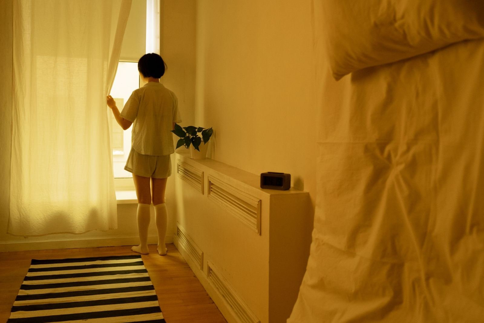 In a bright, simple bedroom, someone in pajamas pulls back a white curtain.