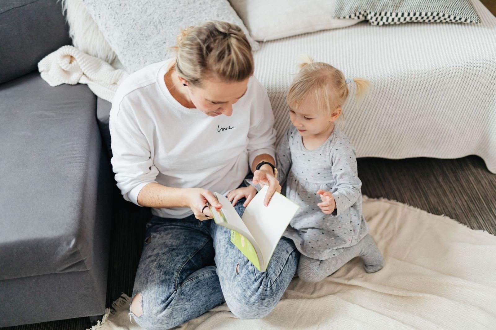 A mother and her small child sit on a rug, reading a book side by side.
