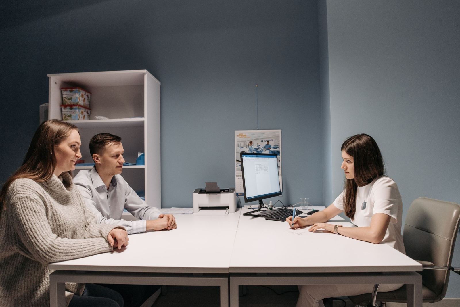 A healthcare worker in white scrubs talks to a couple at a desk
