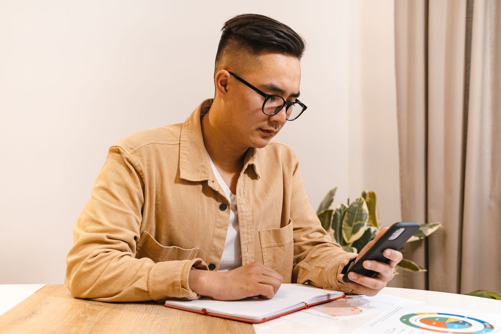 A man with glasses looks at his phone while sitting at a table with a notebook