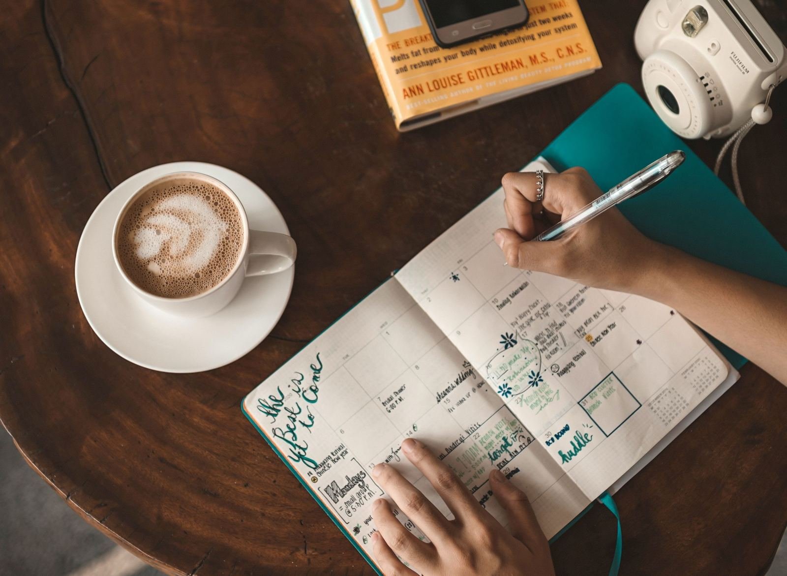 Someone fills out a weekly planner while sitting near a book and a cup of coffee.