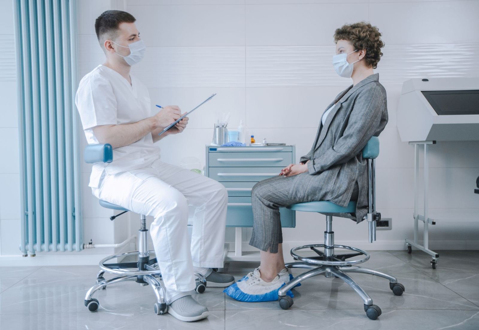 A masked patient in a suit talks with a masked doctor in scrubs at a clinic.