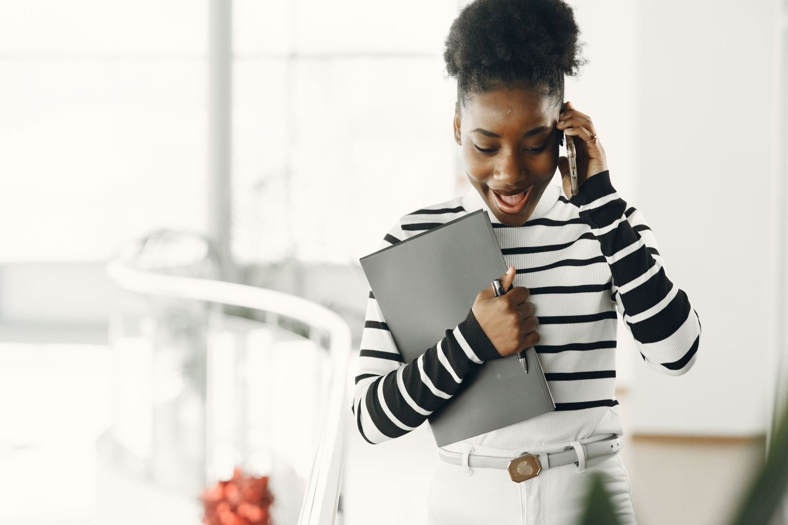 A smiling woman in a striped shirt talks on a phone while holding a folder