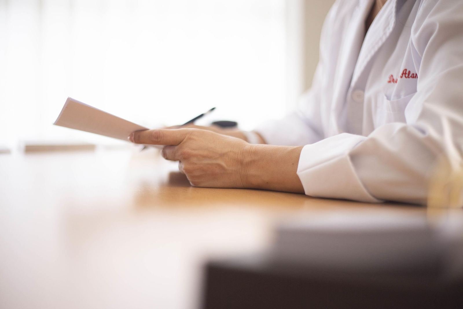A person in a white lab coat holds a small card and pen at a wooden desk