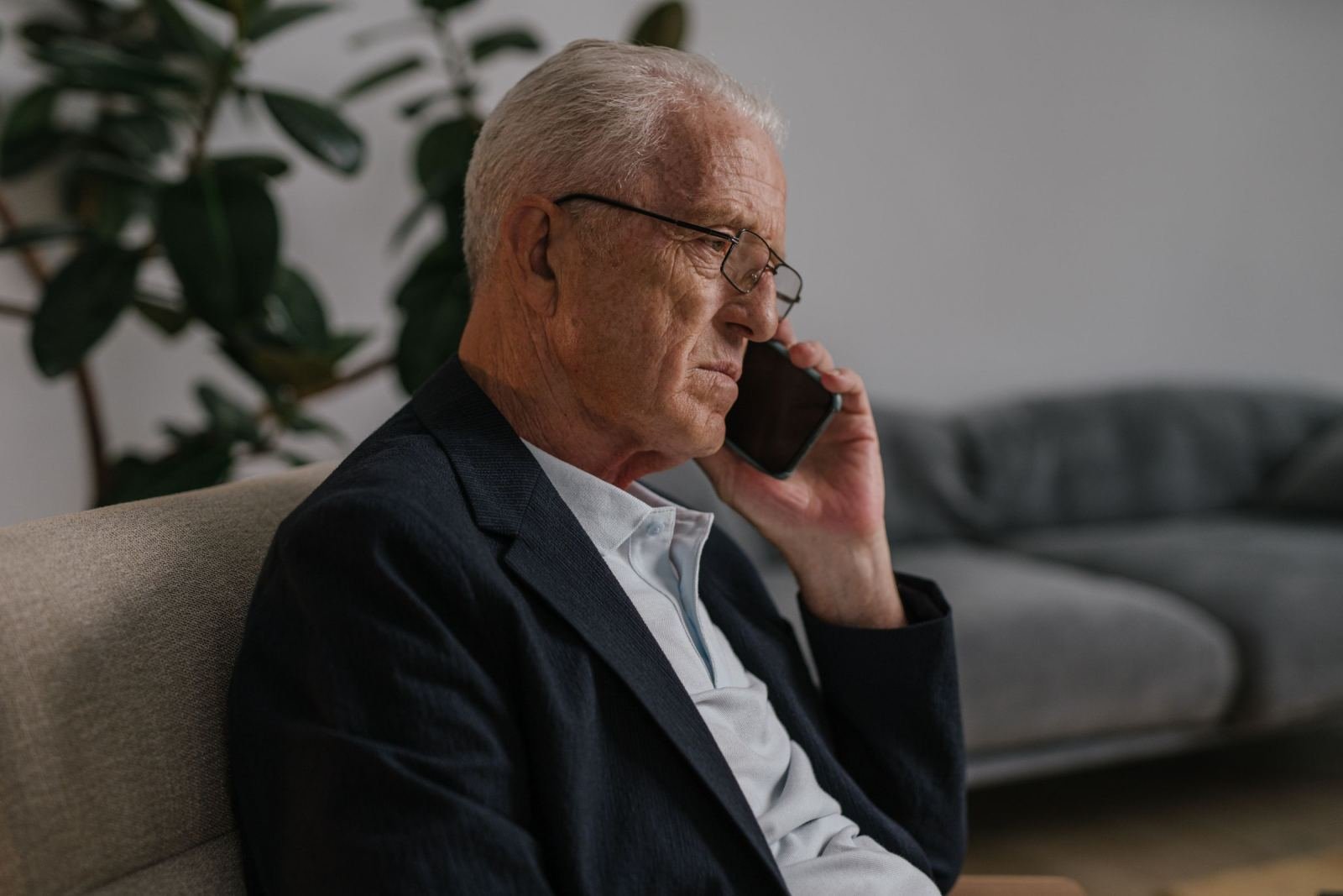 A senior man in a jacket talks on his cell phone from a sofa.