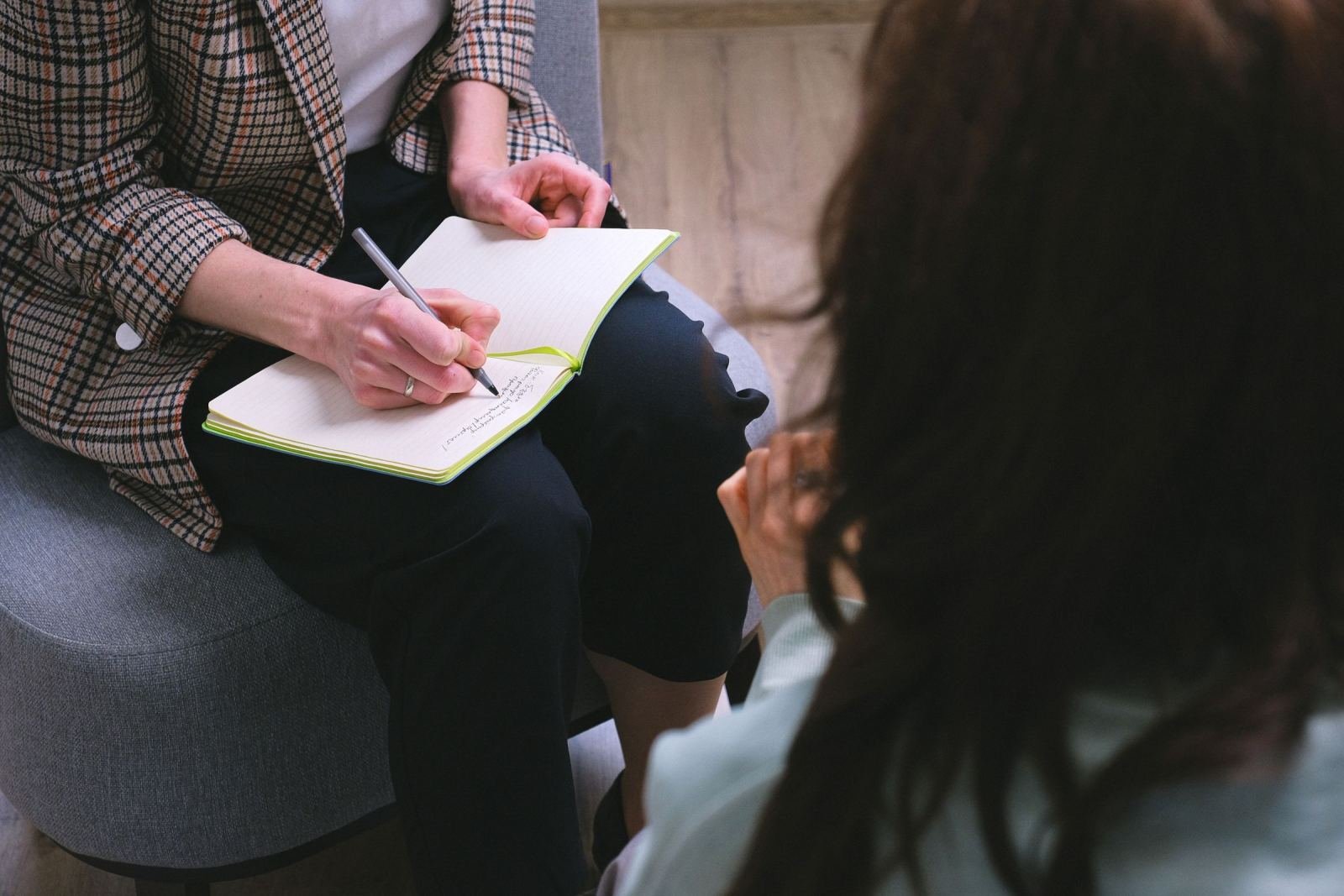 A person in a plaid blazer writes in a notebook while talking with a client