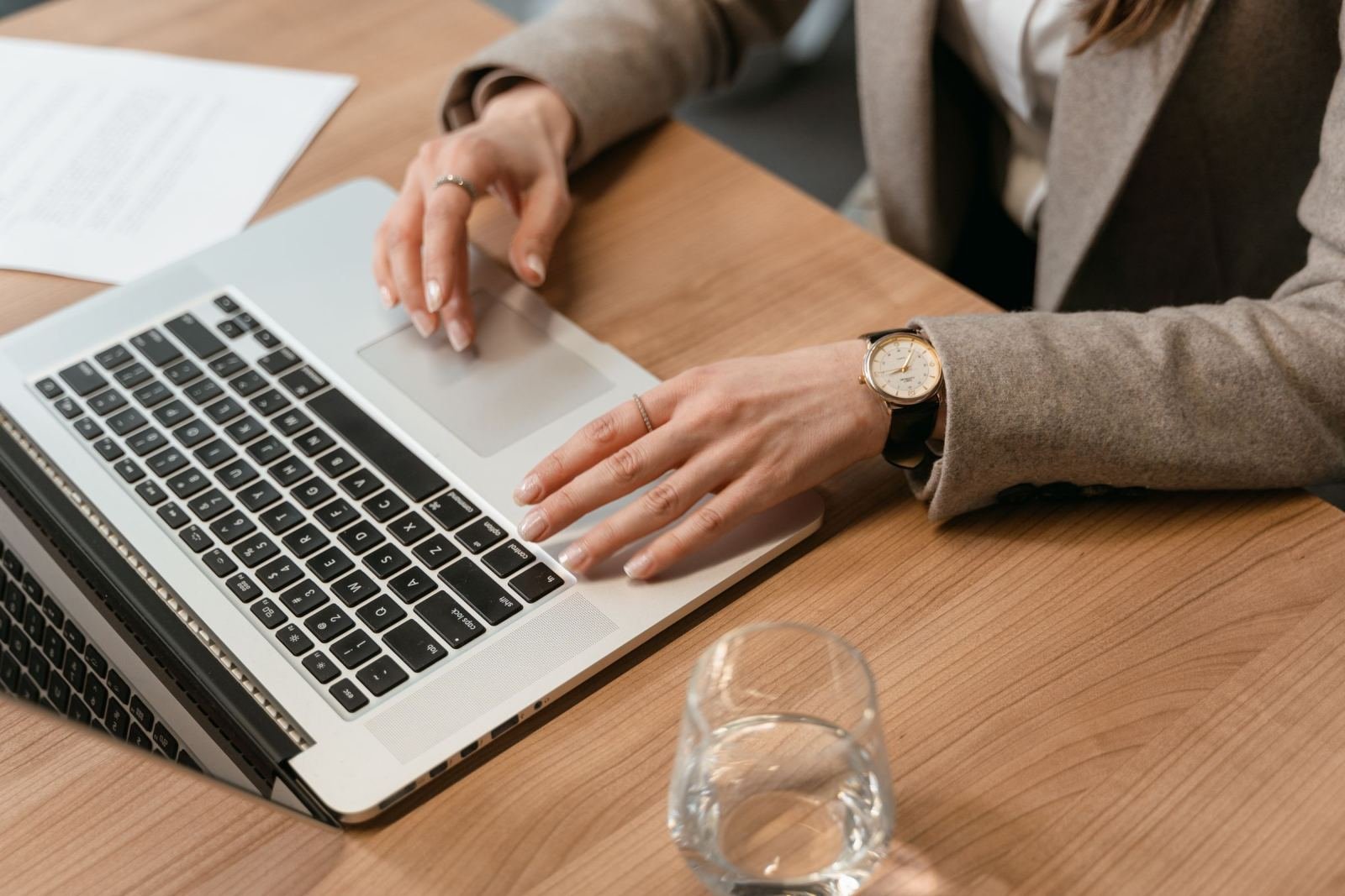 A person in a blazer uses a laptop at a wooden desk with a glass of water