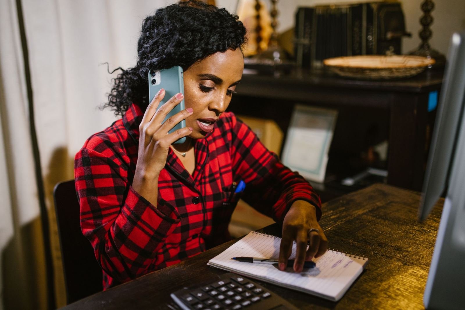 A woman chats on her phone while jotting notes at a desk.