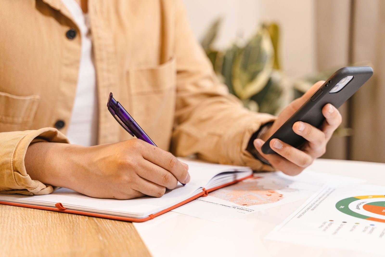 A person writing in a notebook while holding a smartphone at a table