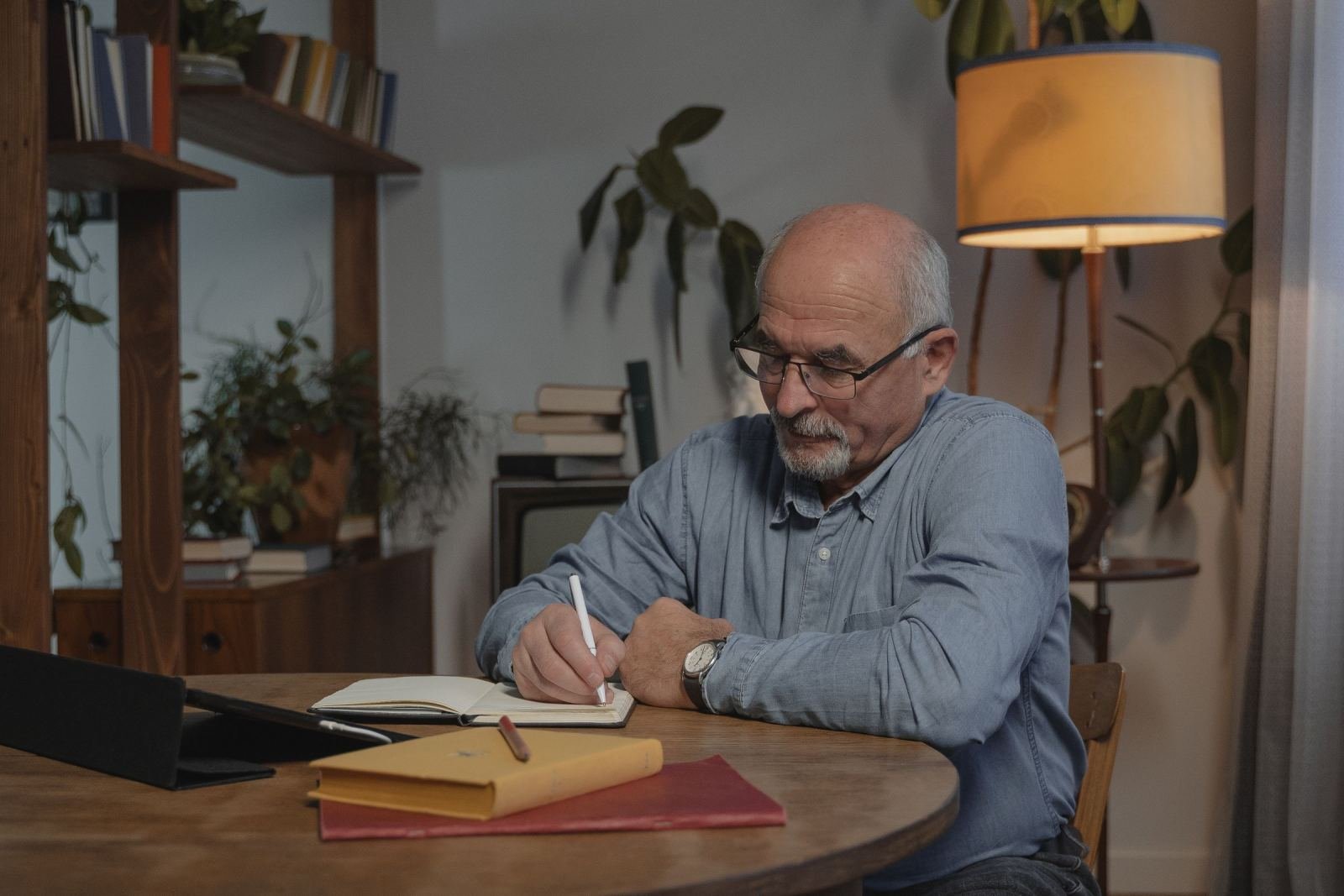 A senior man wearing glasses sits at a wood table writing in a notebook