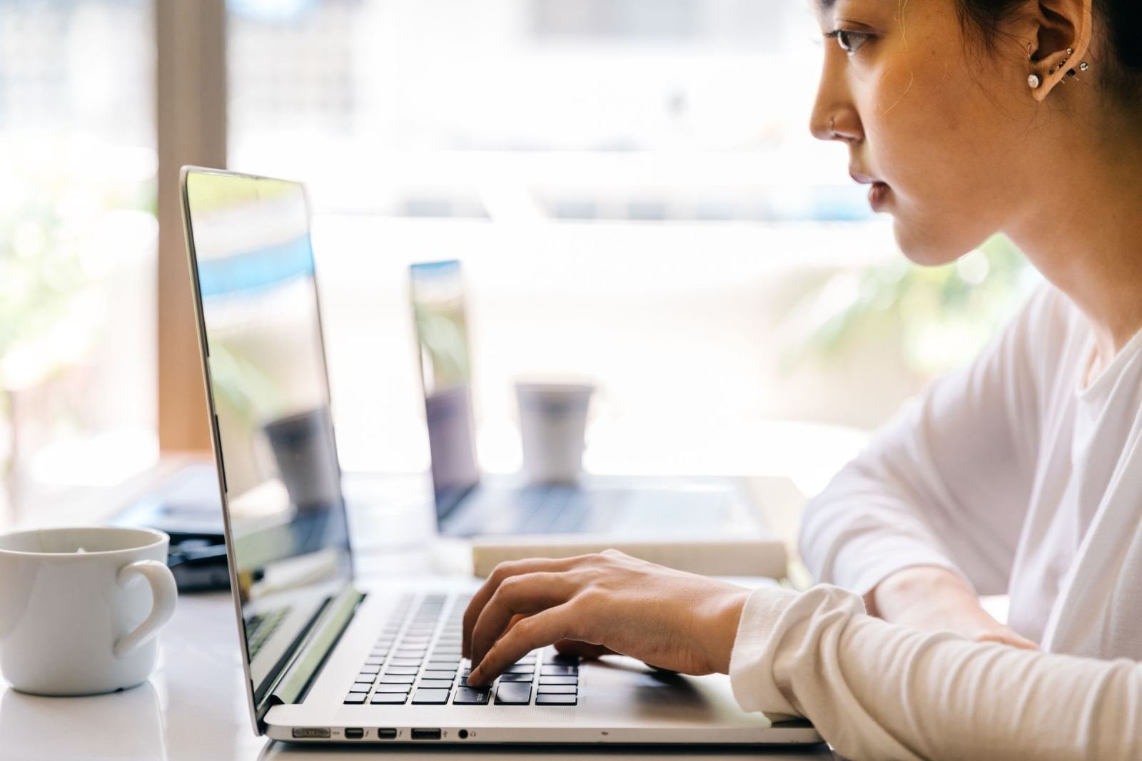 A woman sits at a bright desk, typing on a laptop. A white mug rests near her.
