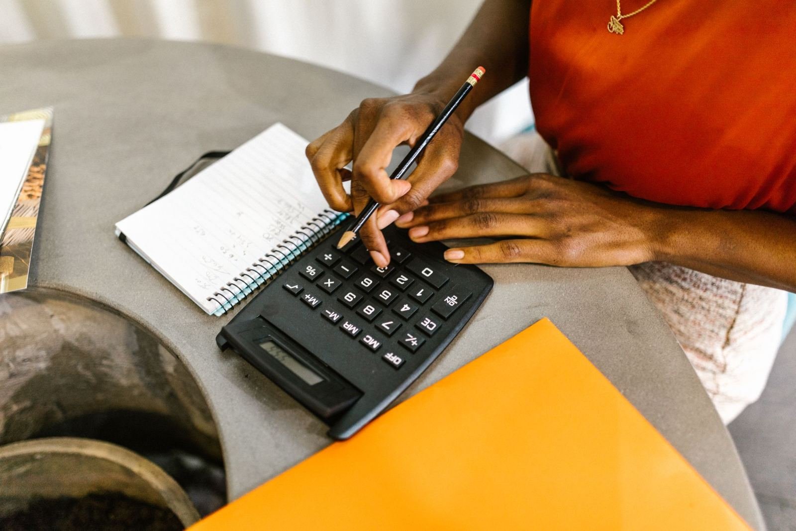 A person using a calculator next to a notebook and an orange folder