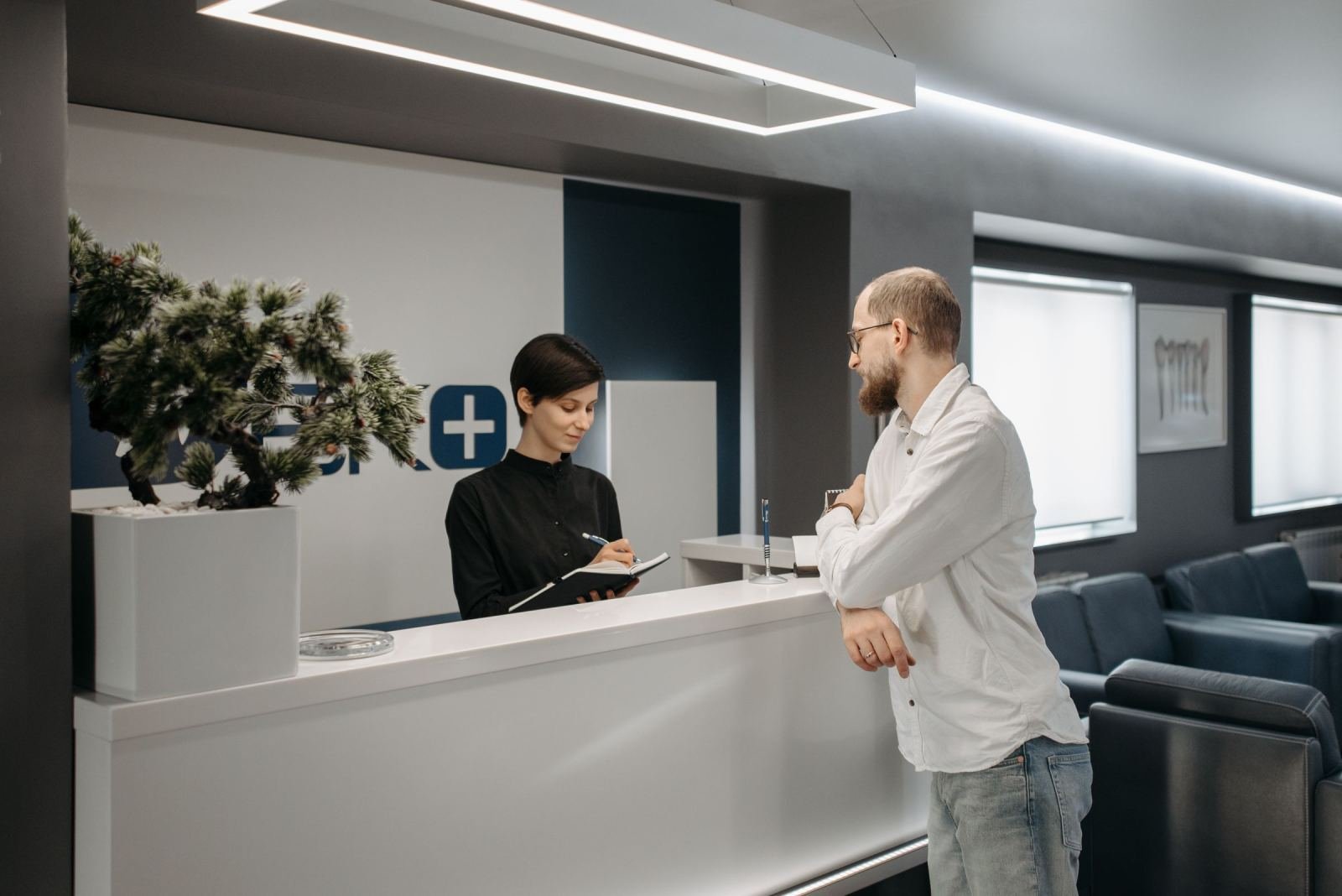 A woman behind a reception desk writes in a notebook while talking to a man